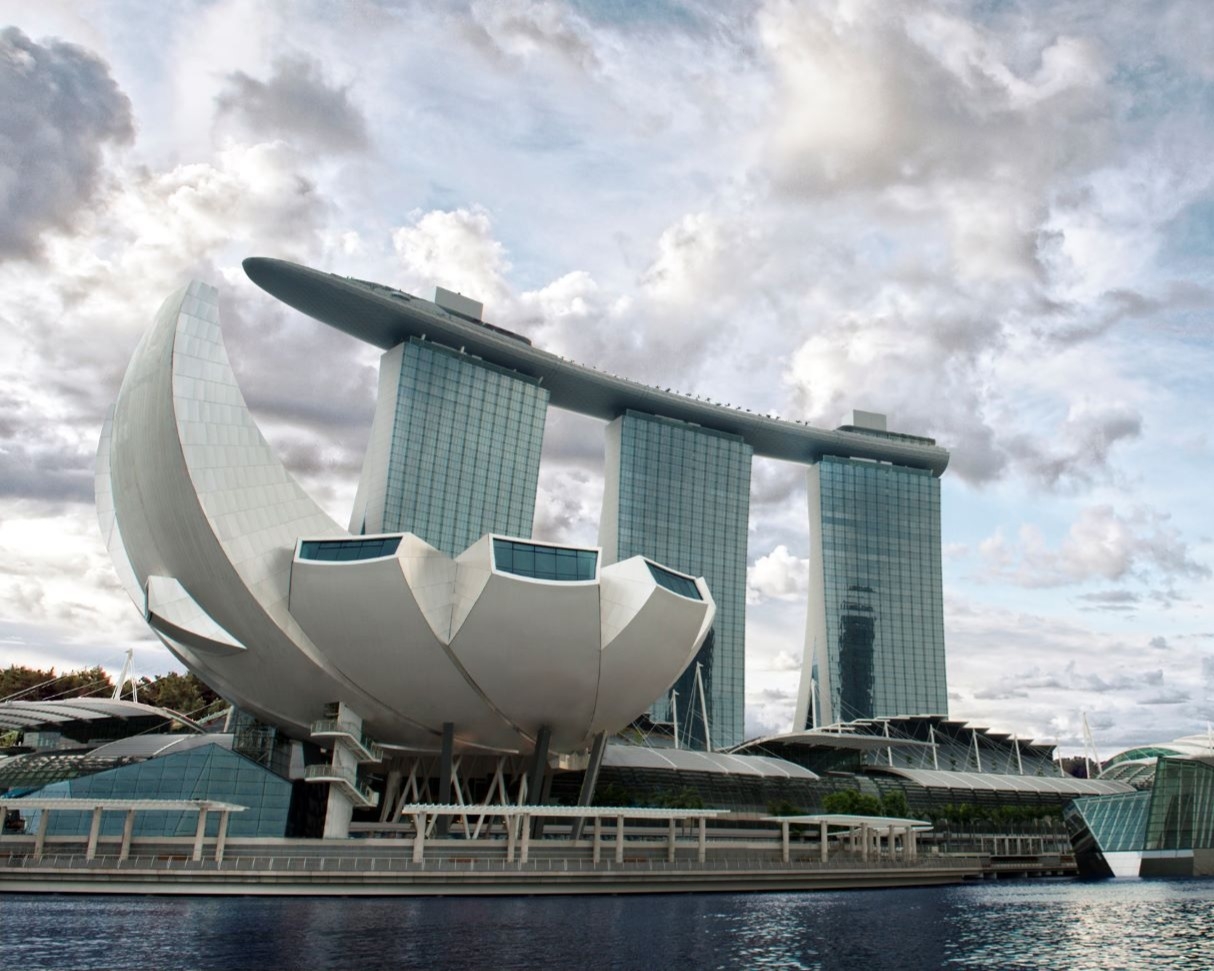 Marina Bay Sands in Singapore featuring three tall glass towers topped by a boat-shaped SkyPark, with the white lotus-shaped ArtScience Museum in the foreground under a cloudy sky.