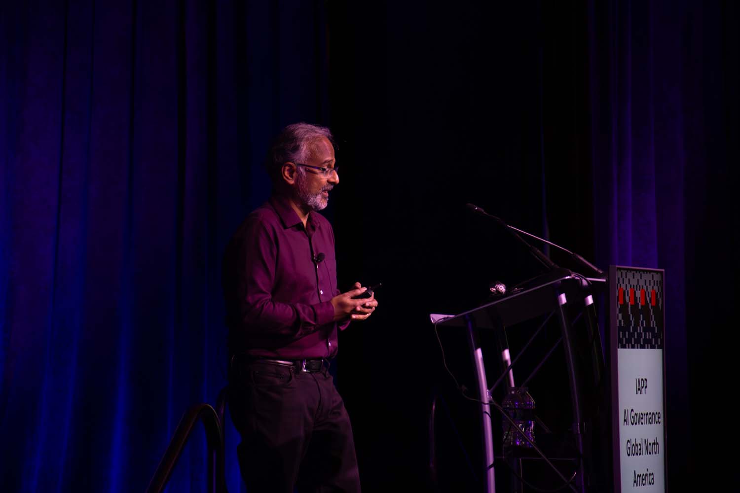 A keynote speaker walks across a wide stage as large presentation screens display “Chris Miller” and session details, all illuminated in purple and blue conference lighting.