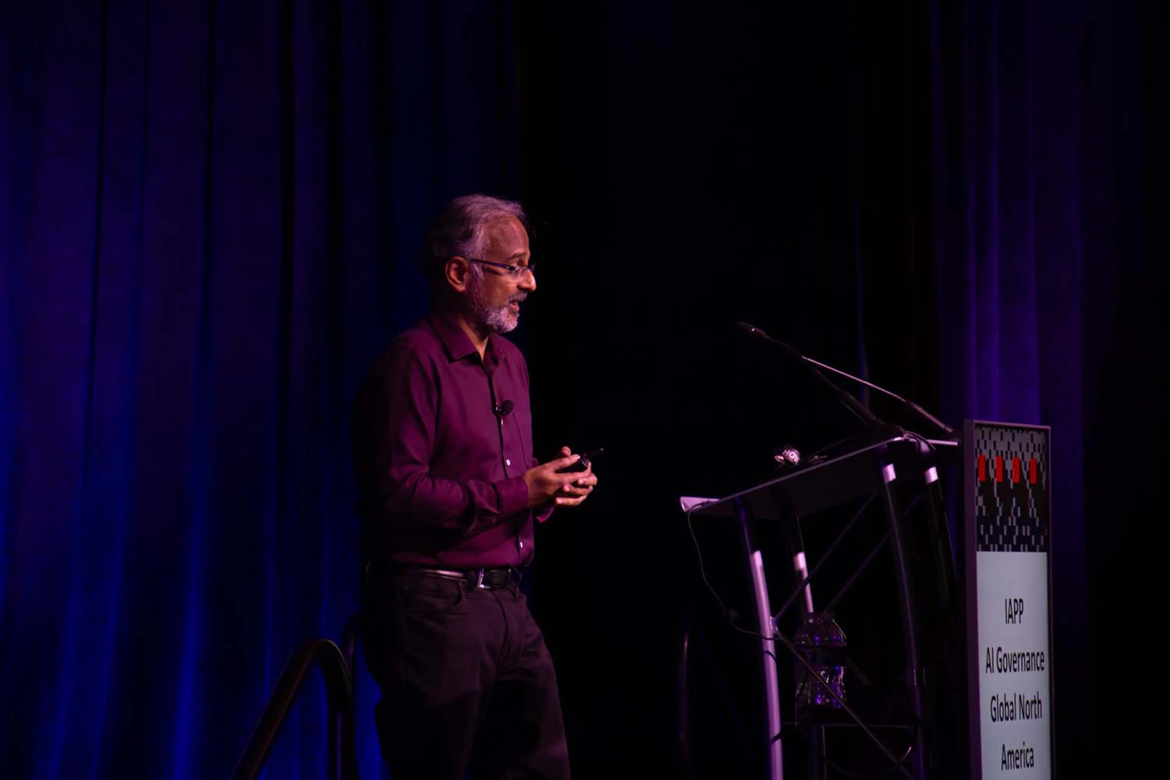 A keynote speaker walks across a wide stage as large presentation screens display “Chris Miller” and session details, all illuminated in purple and blue conference lighting.