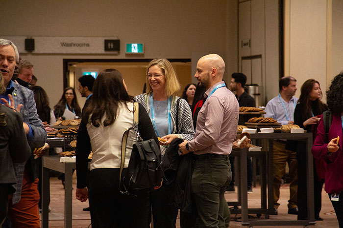 Conference attendees gathered around high tables during a networking break, holding drinks and food while conversing in a hallway with signage for restrooms in the background.