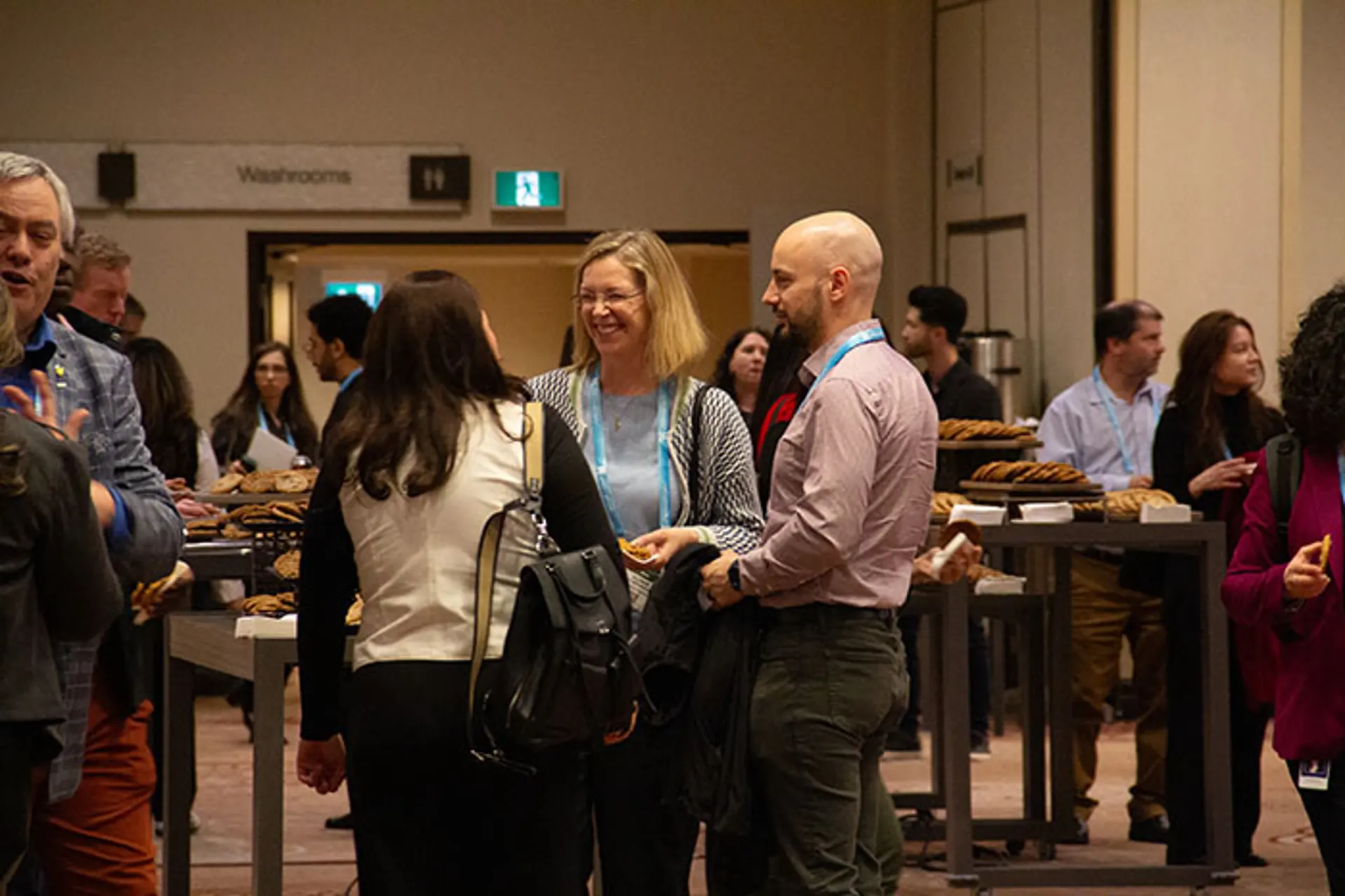 Conference attendees gathered around high tables during a networking break, holding drinks and food while conversing in a hallway with signage for restrooms in the background.