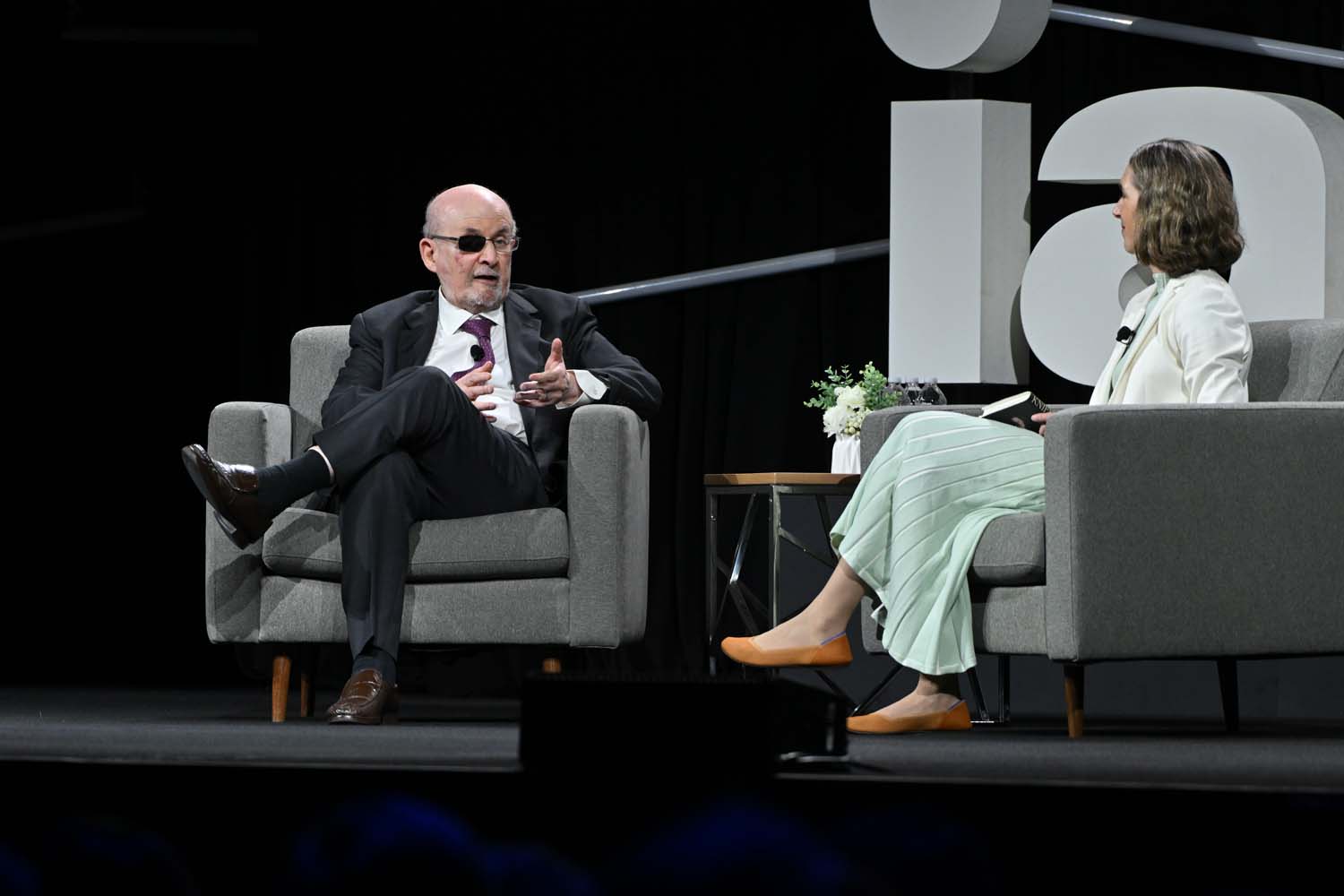 A close-up view captures a seated speaker gesturing while speaking, with the other speaker listening nearby and large IAPP letters looming behind on the darkened stage.