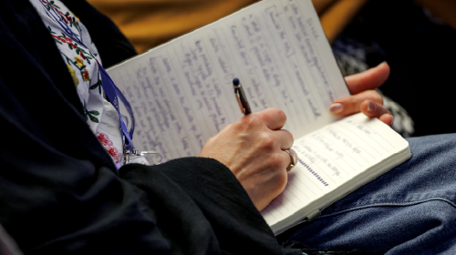 Close up of a person writing in a notebook. The notebook is full of text and the person has a pen in hand.
