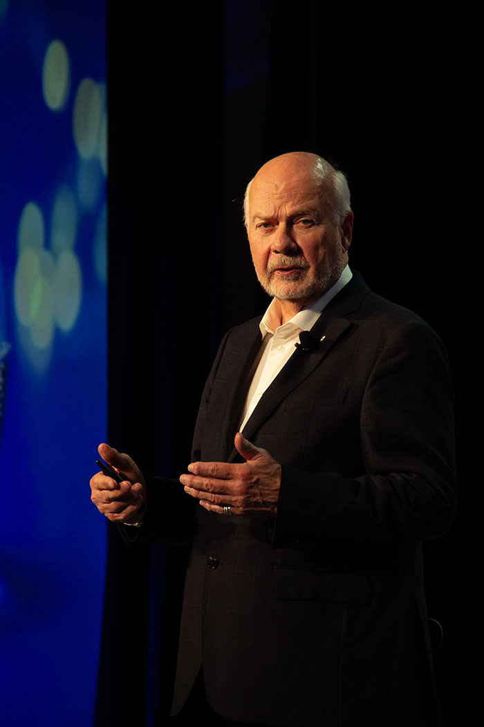 Speaker presenting on stage in a dark conference setting, holding a remote and gesturing with hands, with a blue-lit backdrop and abstract light patterns.