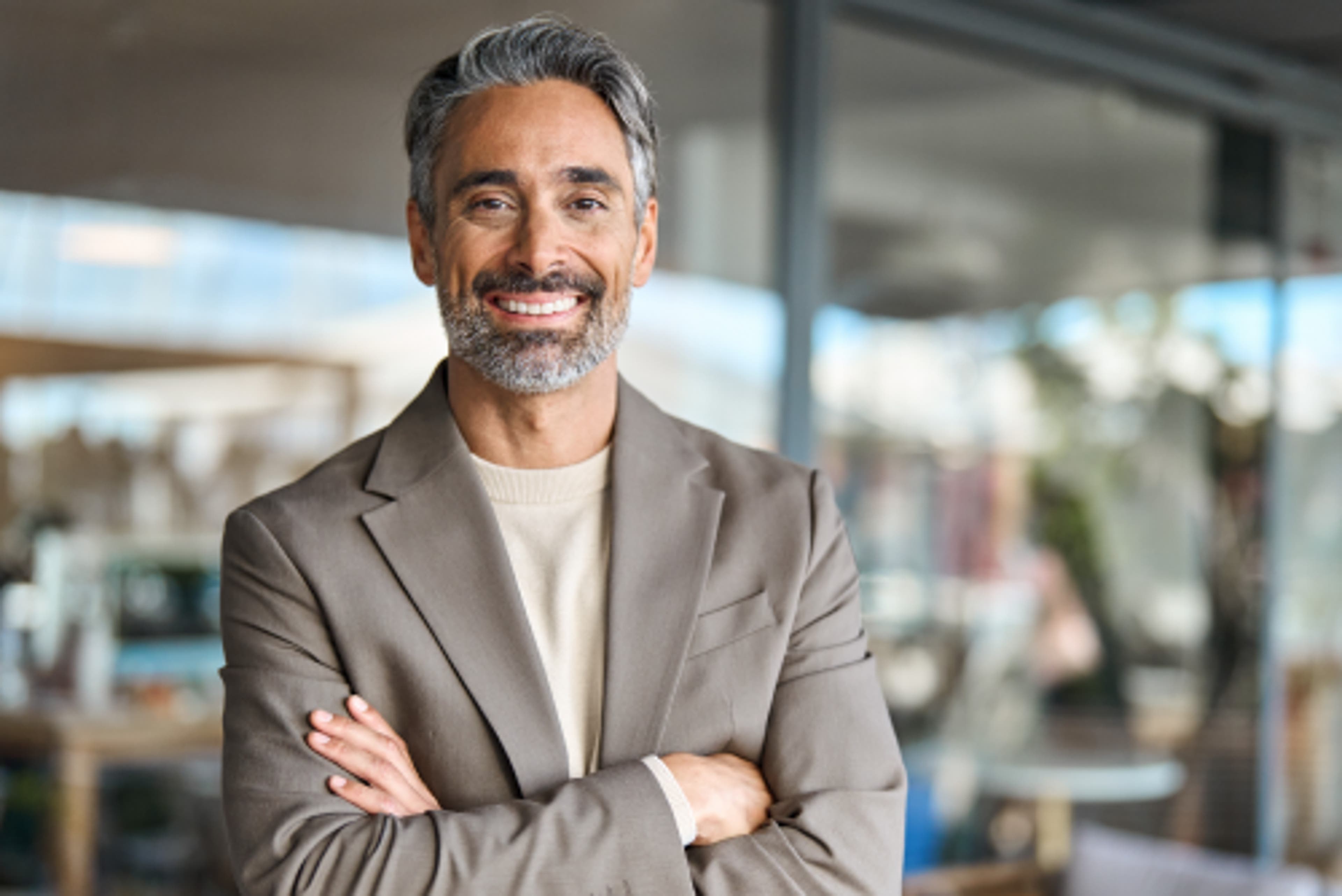 A man with partially gray hair and a beard wearing a sport coat with his arms crossed. He is smiling.