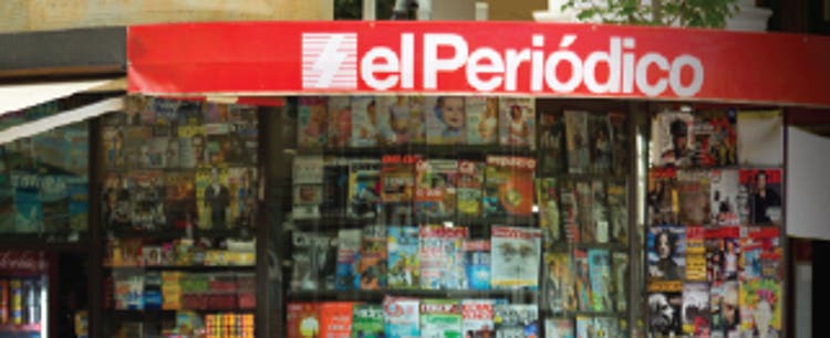 An image of a Spanish newspaper stand, reading el Periódico in white lettering against a red background. A variety of newspapers and magazines fill the display racks below the signage.