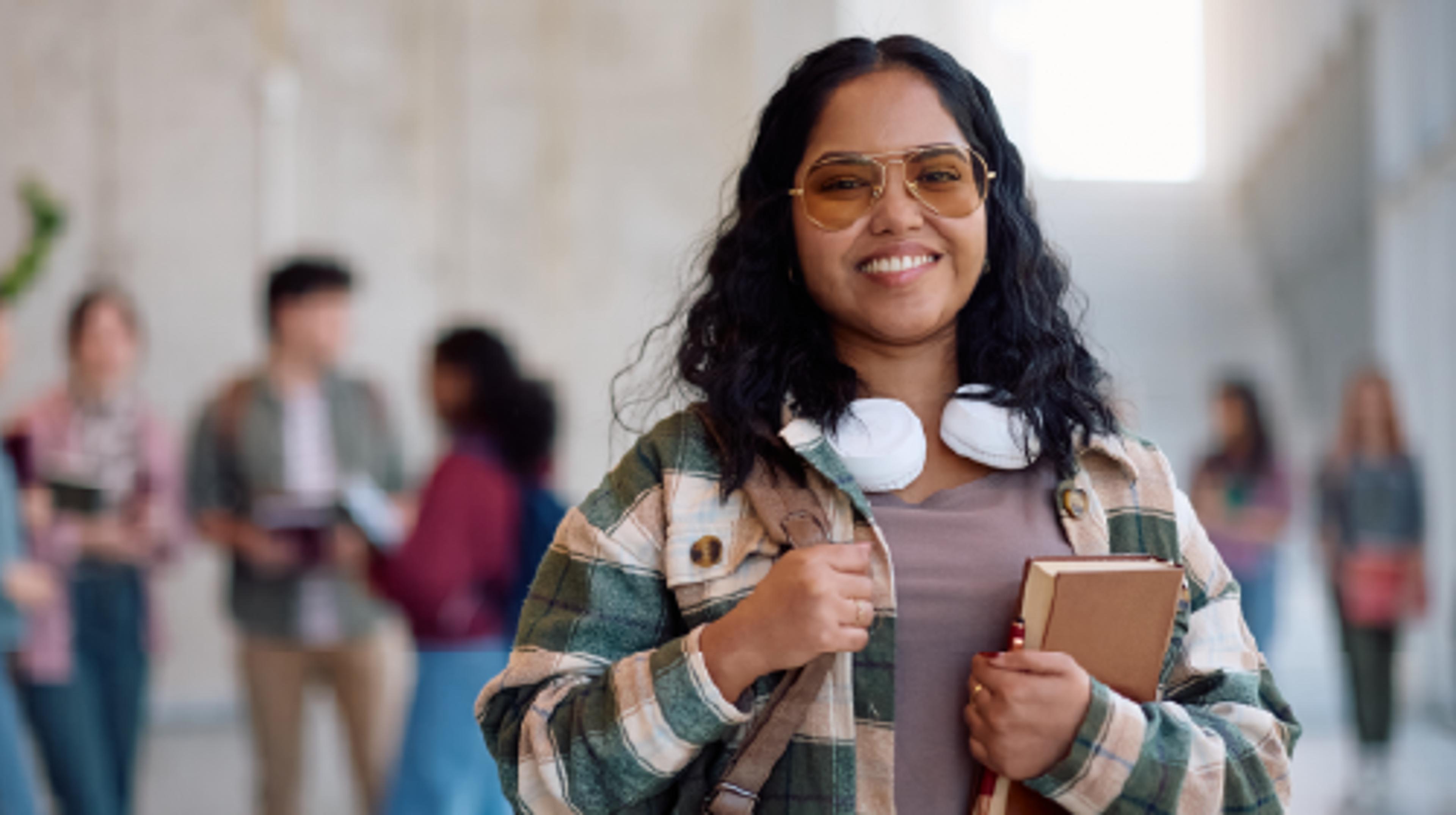 A young woman wearing a flannel shirt and dark glasses with headphones around her neck is facing the camera and smiling
