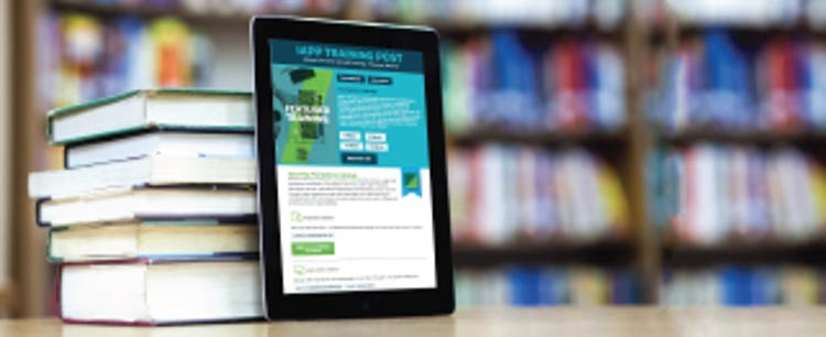 An iPad rests upright on a flat, low-sheen wood surface against a stack of six books of varying sizes and heft. In the background are a collection of out-of-focus bookshelves, filled with books of varying colors. The iPad screen is on, displaying a sample of IAPP's Training Post content.