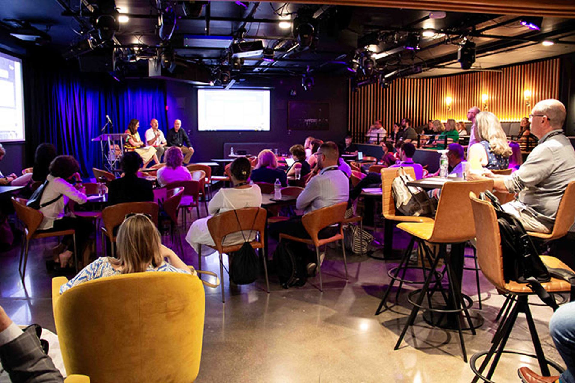 Conference session in a dimly lit venue with stage lighting, featuring a panel discussion at the front and attendees seated at tables and high chairs.