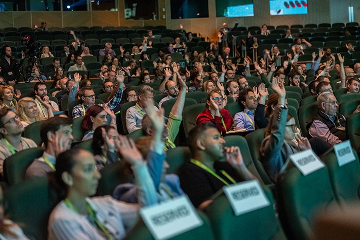 Audience seated in an auditorium with many raising hands during an interactive session. Reserved signs are visible on some seats.
