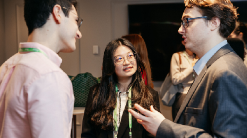 A young woman in glasses is in the center of the photo. She is looking at the man to her right, who is speaking; another man on the left is listening and smiling.