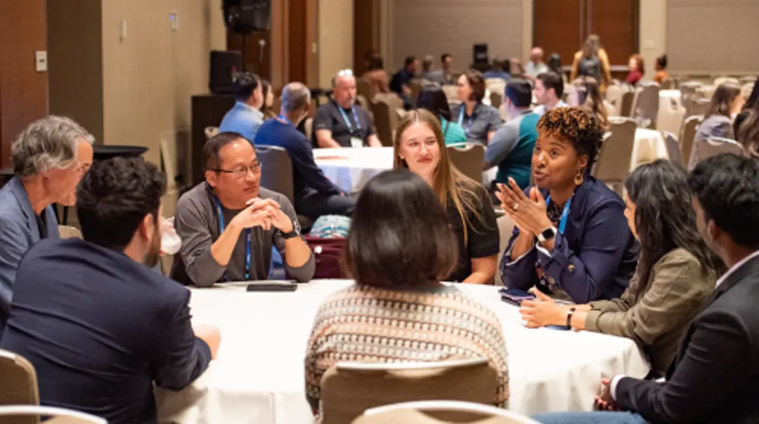 A group of people seated around a round table at a conference, engaged in discussion, with additional attendees and tables visible in a hotel meeting room in the background.