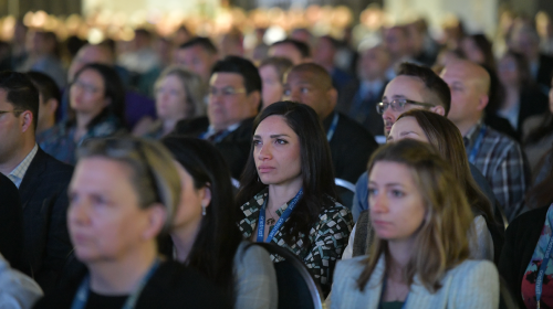 Close-up view of conference attendees seated in rows, listening to a presentation, with a large audience visible in the background.