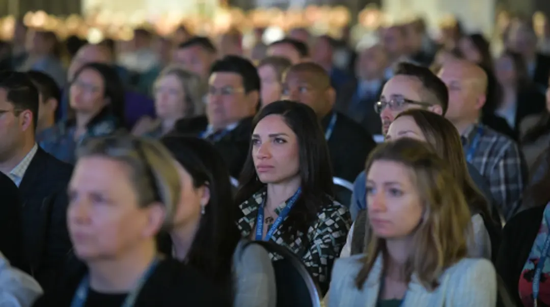 Close-up view of conference attendees seated in rows, listening to a presentation, with a large audience visible in the background.