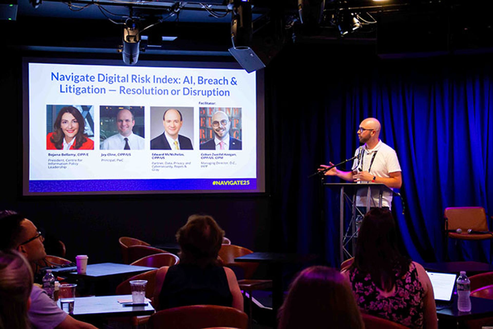 Speaker presenting at a podium in a dark room with blue stage lighting. A large screen behind displays “Navigate Digital Risk Index: AI, Breach & Litigation — Resolution or Disruption.”