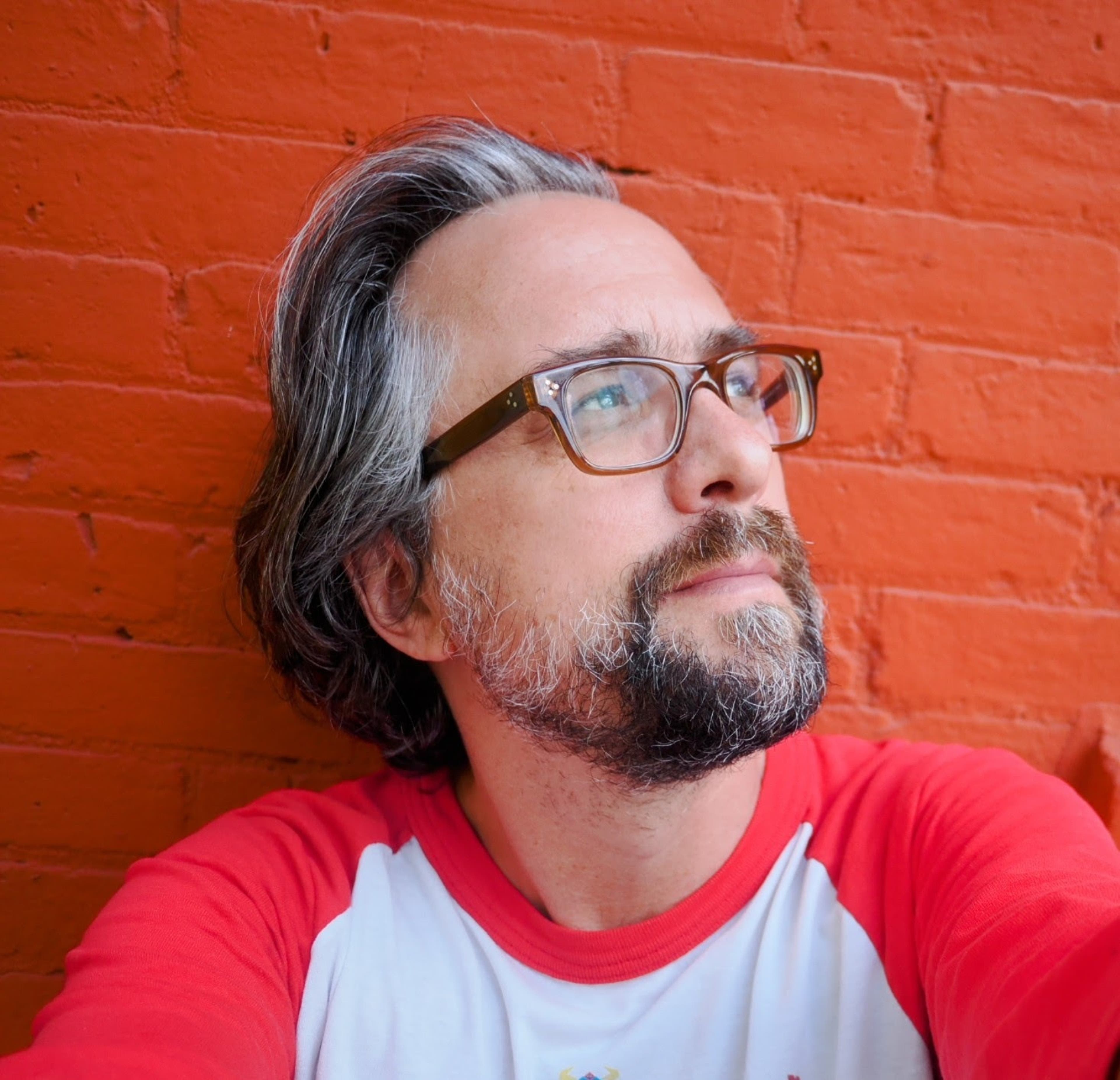 Artist Jer Thorp; he has a beard and glasses and is standing with his arms crossed looking at the camera.