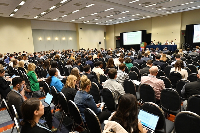 Large audience seated in rows of chairs facing a panel discussion at the front of a conference room. A green event sign is visible near the stage, and many attendees have laptops or notebooks.