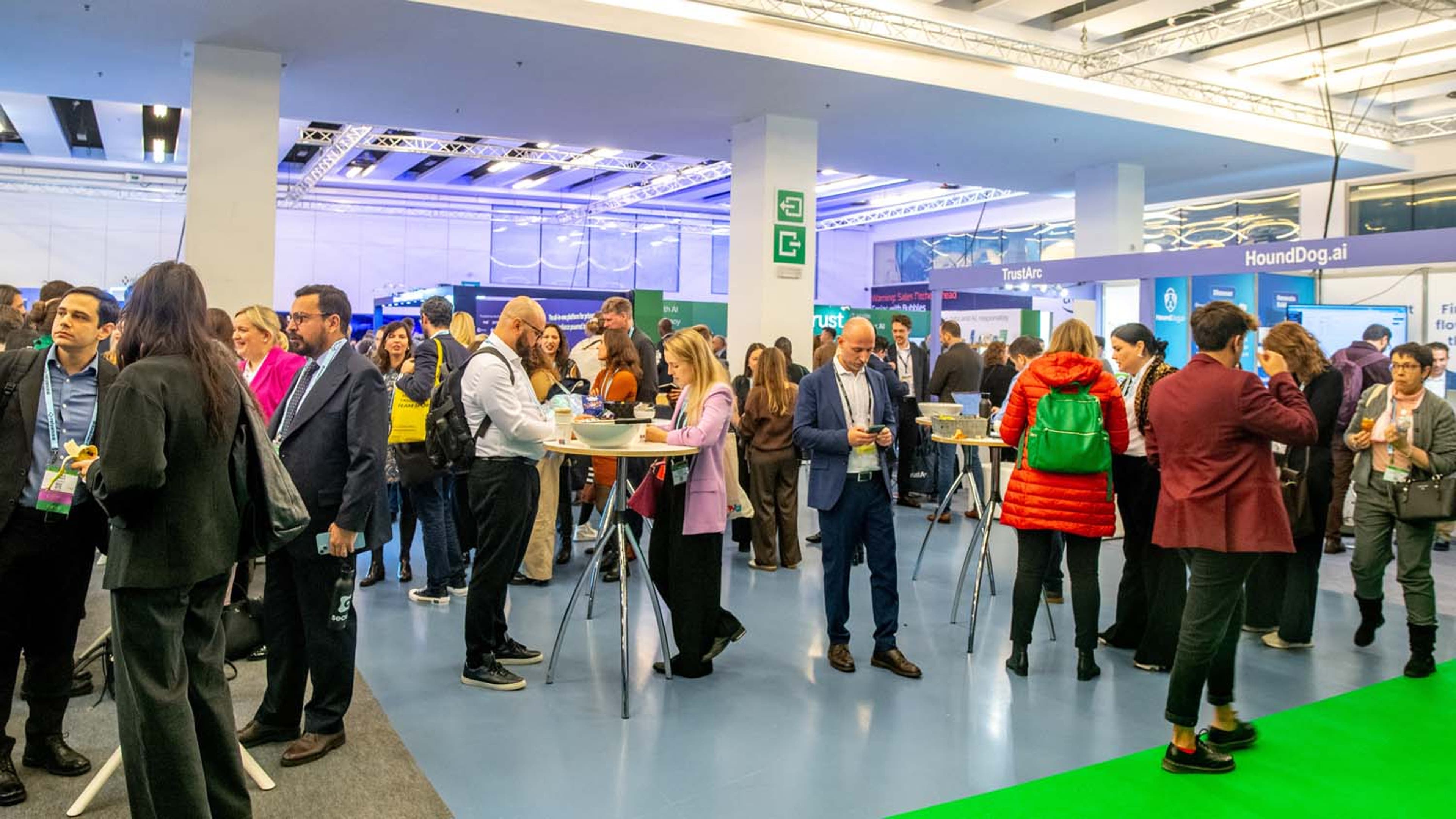 Busy exhibit hall with delegates standing and talking near high tables. Booths with company banners are visible in the background.