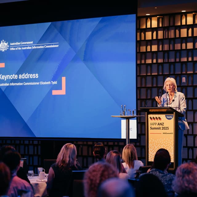 Keynote speaker Elizabeth Tydd stands at podium on stage during IAPP ANZ Summit 2025. A large screen showing a blue background and the words "Keynote address, Australian Information Commissioner Elizabeth Tydd" is visible behind her. A crowd is visible in front of her.