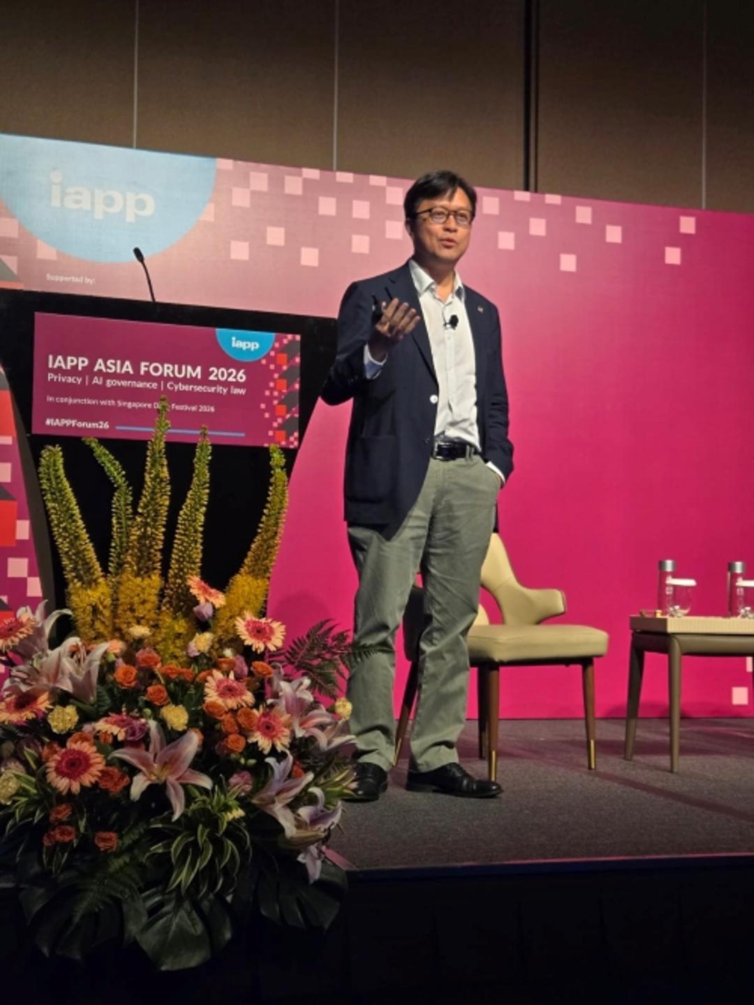 A speaker stands on stage at the IAPP Asia Forum 2026, delivering a presentation beside a podium and a large floral arrangement. A pink conference backdrop displays event branding, and two chairs with a small table are positioned on the stage behind the speaker.