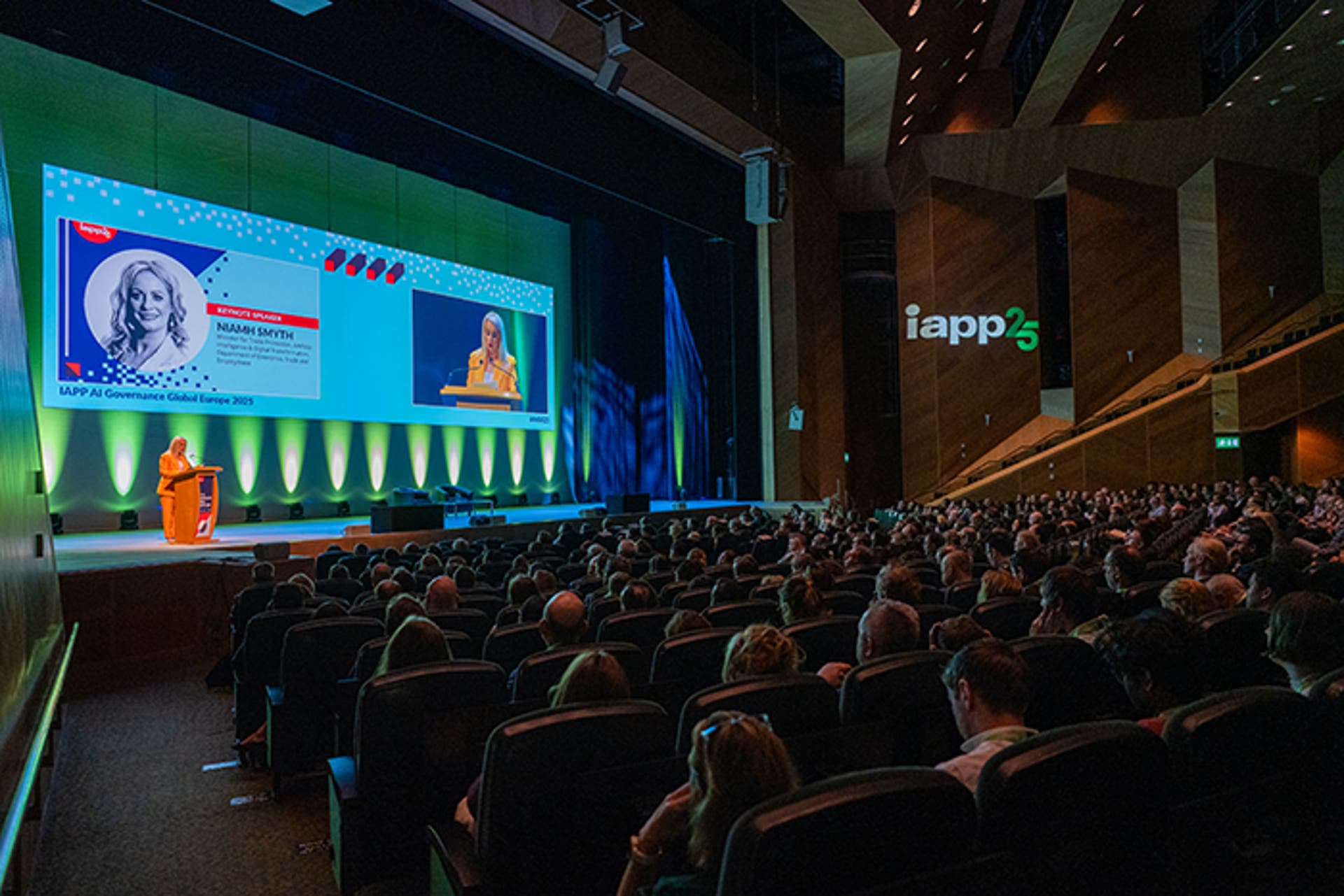 Conference keynote session in a large auditorium. A speaker stands at a podium on stage with a big screen showing name and event branding. Audience fills the seats.