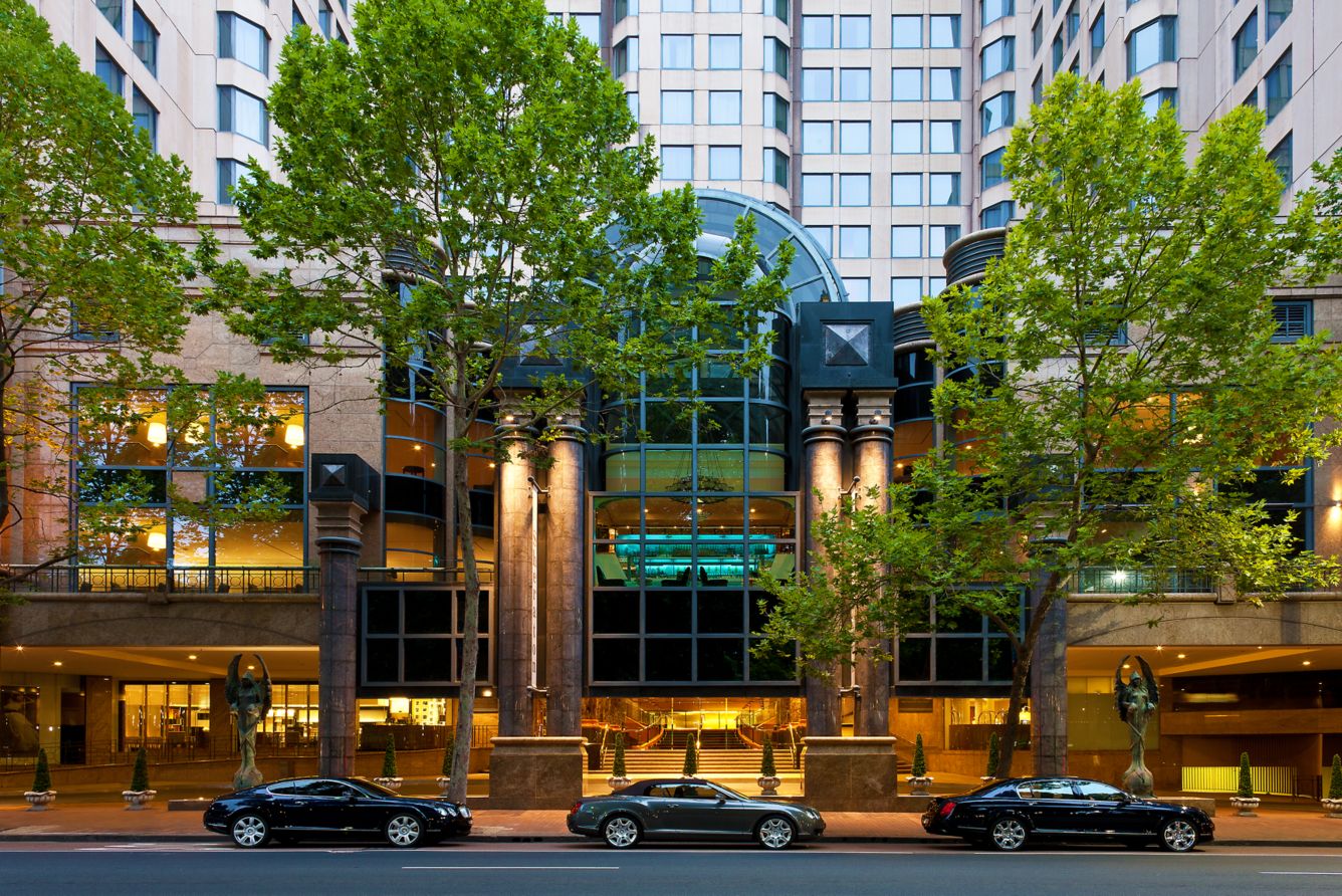 Exterior view of a tall hotel building with a modern glass entrance framed by large stone columns. Two leafy trees partially obscure the facade, and three black cars are parked along the street in front. Warm interior lighting is visible through the windows, highlighting the lobby area.