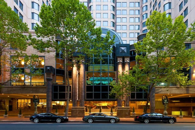 Exterior view of a tall hotel building with a modern glass entrance framed by large stone columns. Two leafy trees partially obscure the facade, and three black cars are parked along the street in front. Warm interior lighting is visible through the windows, highlighting the lobby area.