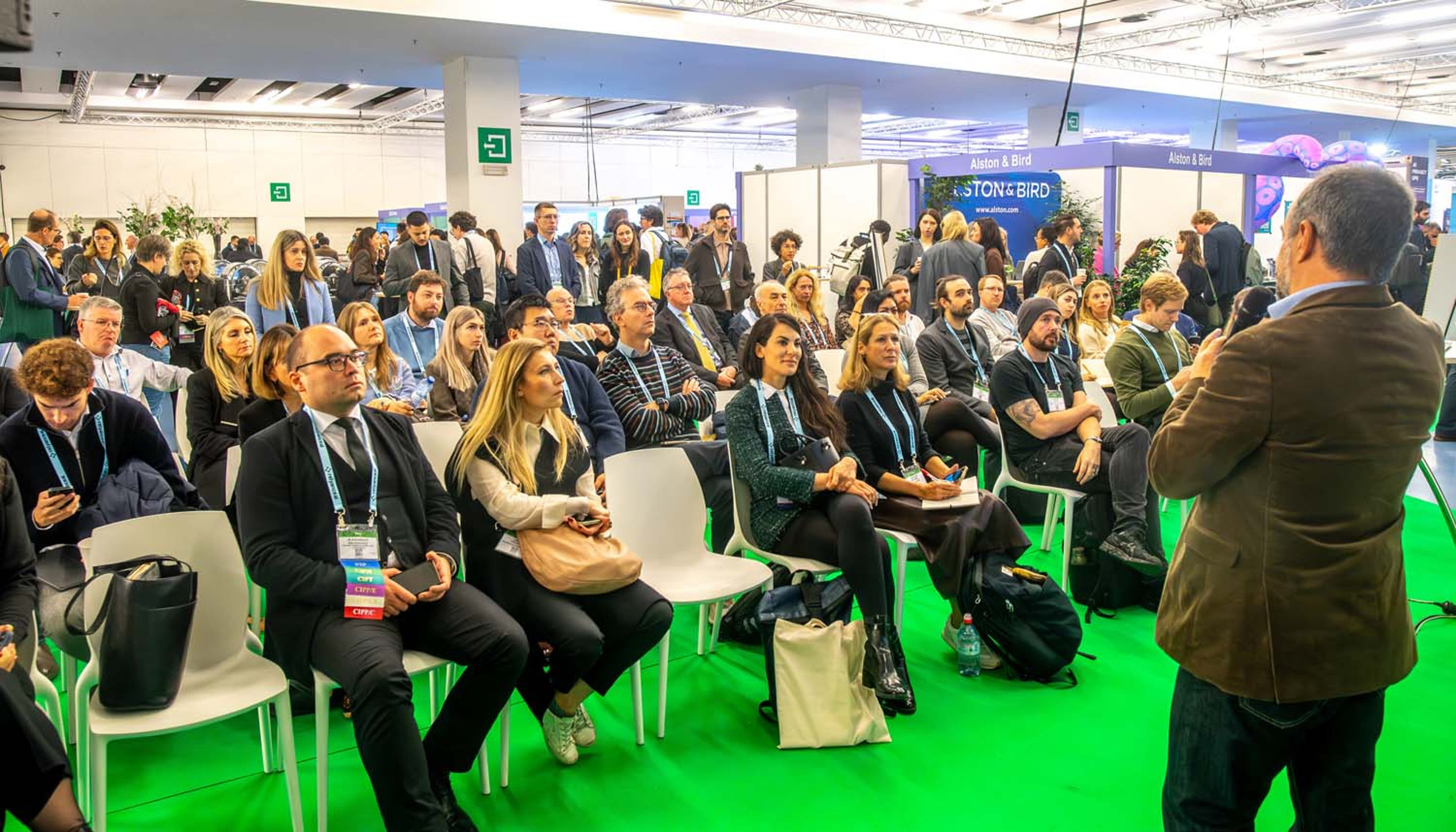 Speaker addressing a seated audience in an exhibit hall area with green flooring. Delegates are listening while others stand near booths in the background.