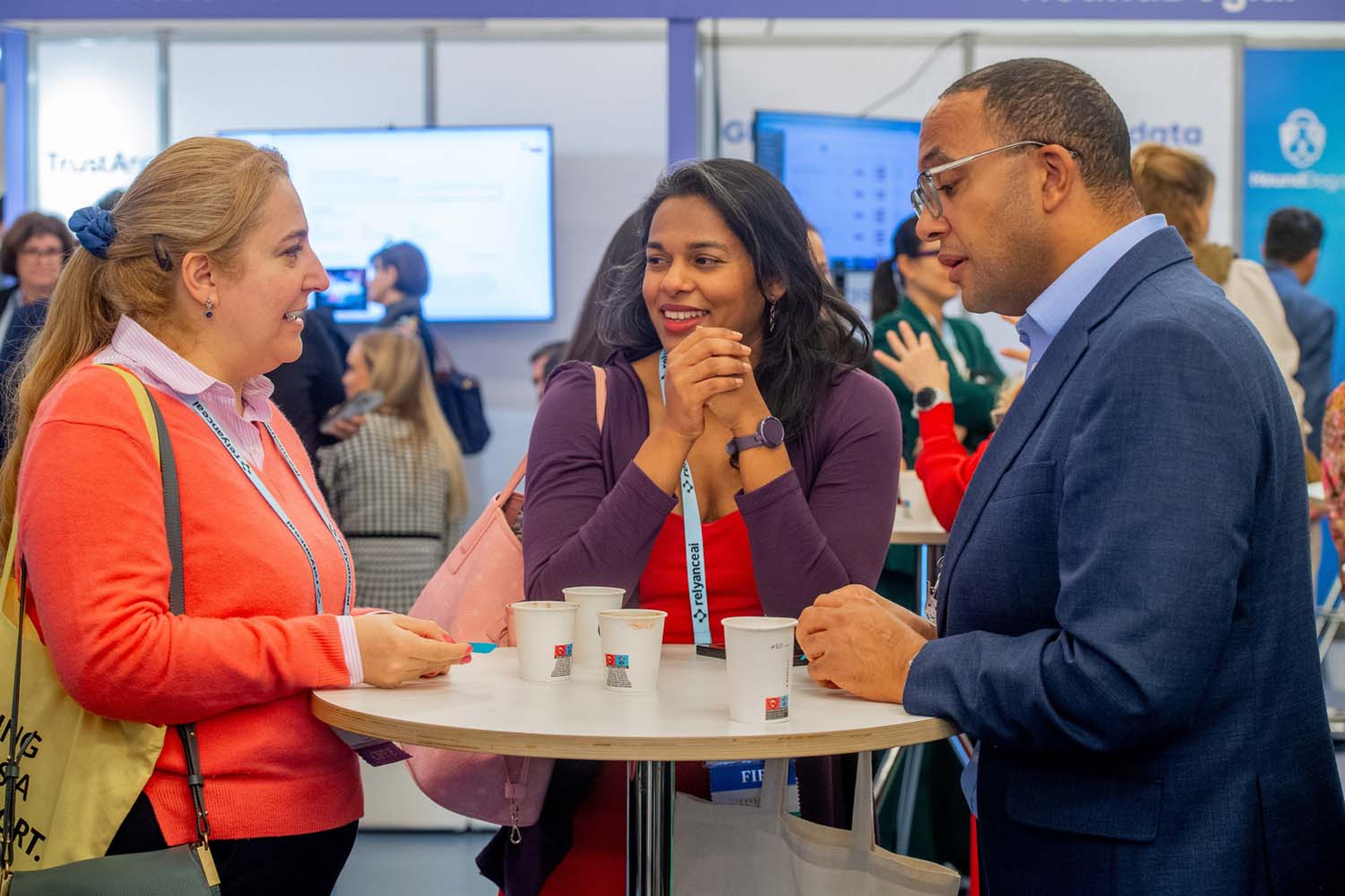 Three delegates standing around a tall round table in an exhibit hall, holding coffee cups and talking. Booths and screens are visible in the background.