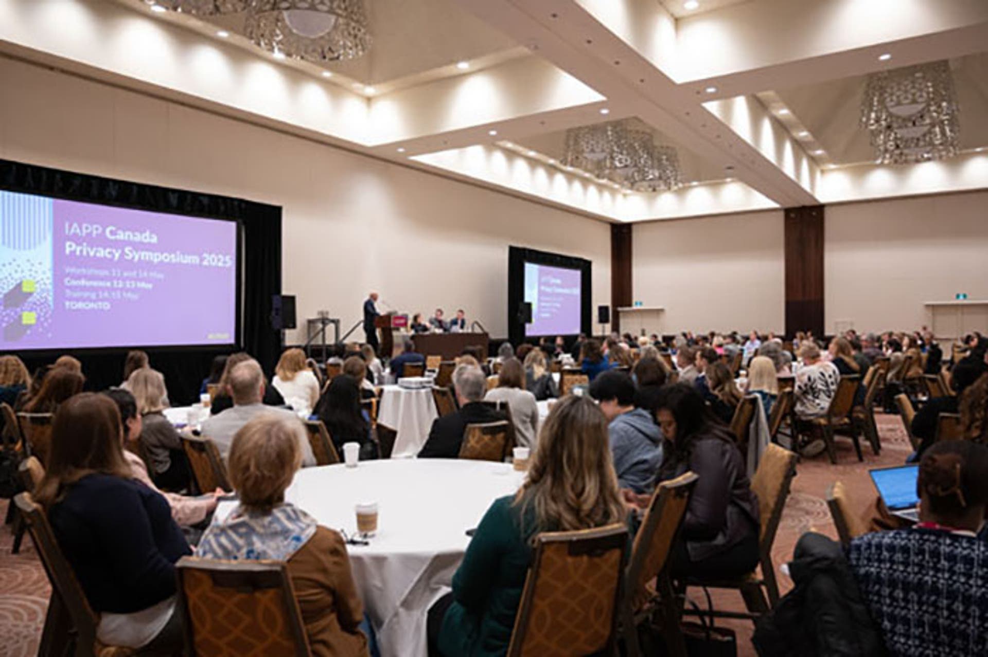 Large conference room with attendees seated at round tables facing a stage. A speaker stands at a podium, and two screens display “IAPP Canada Privacy Symposium 2025” with event details.