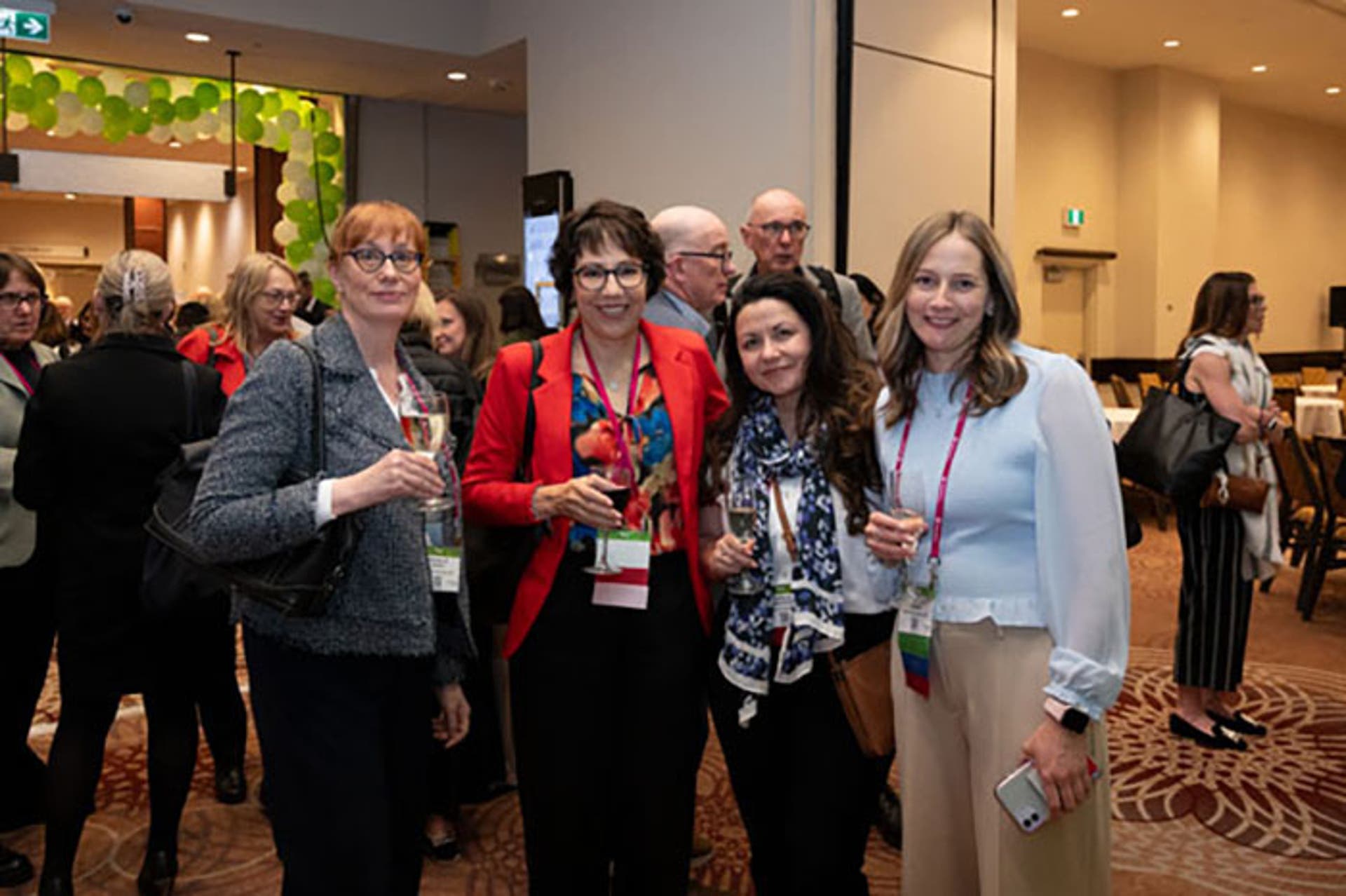 Group of attendees standing together during a networking event, holding drinks and wearing conference badges. Green balloon decorations are visible in the background.
