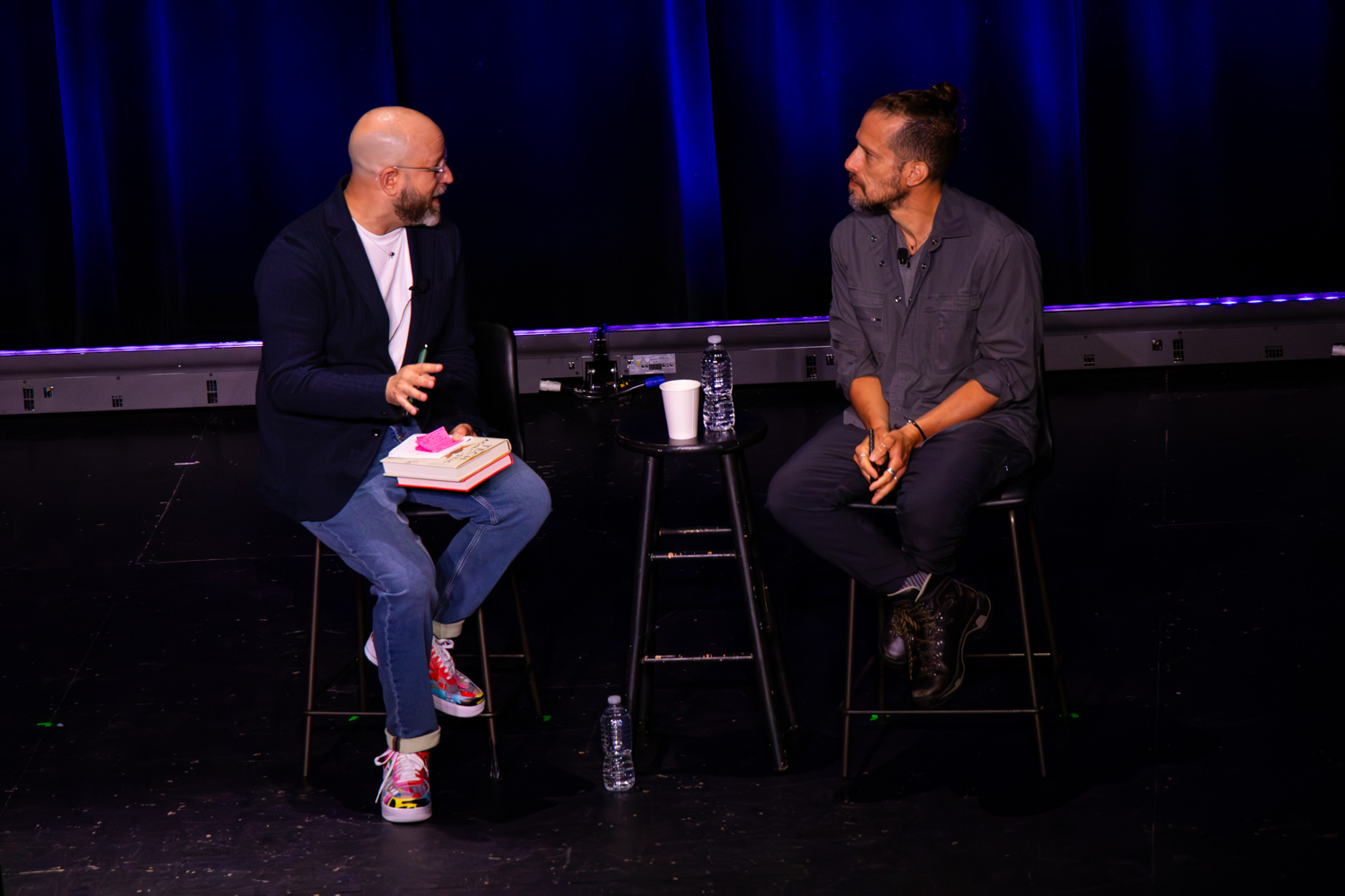 Two speakers sit facing one another on stage in a conversational interview format, with notebooks, water bottles, and a small table between them, framed by deep blue theater curtains and subdued stage lighting.