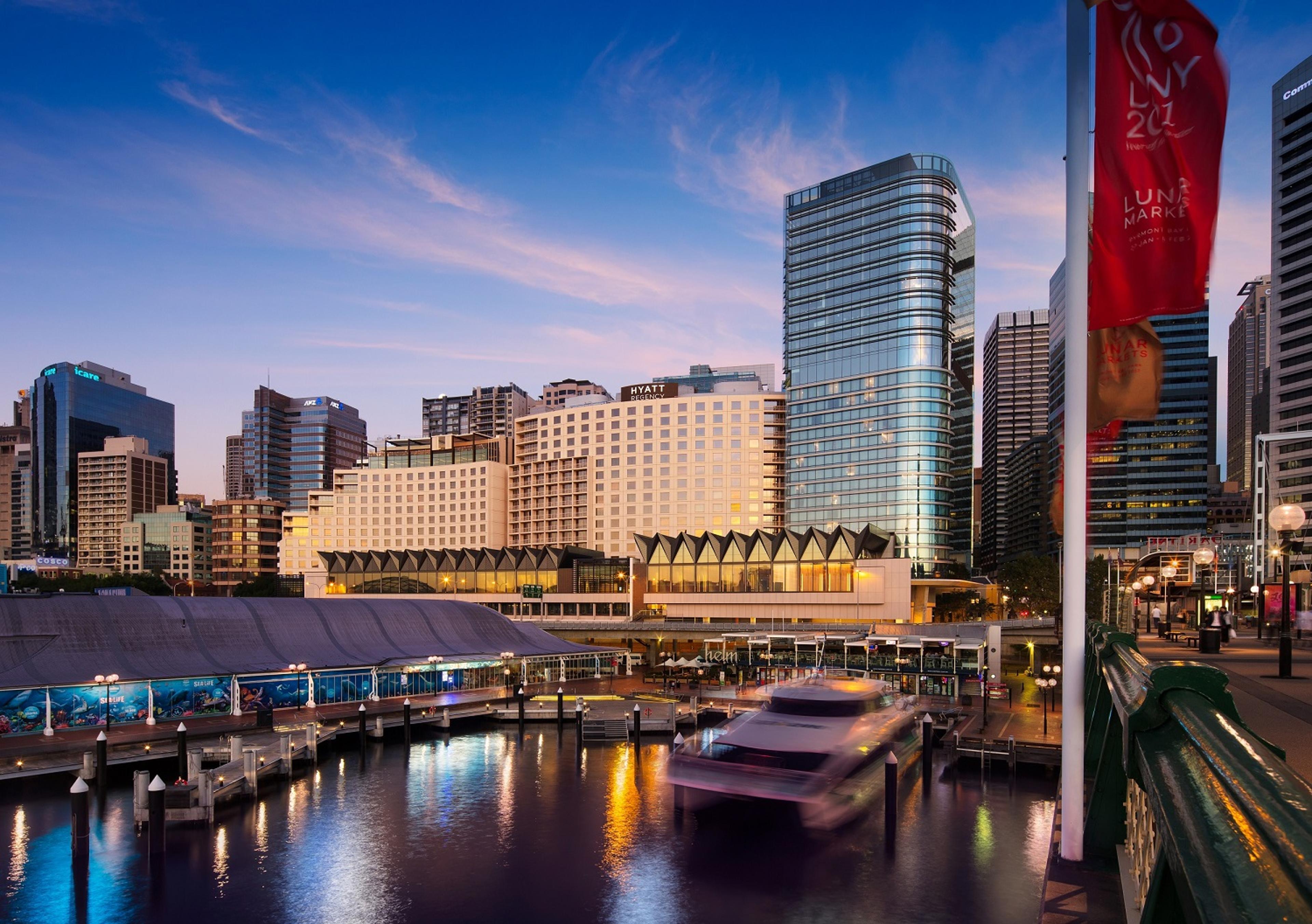 A waterfront city skyline at dusk features modern high-rise buildings and a lit hotel complex reflected in calm water, with a blurring ferry passing through the harbor in the foreground and colorful banners lining a pedestrian bridge under a blue and pink evening sky.