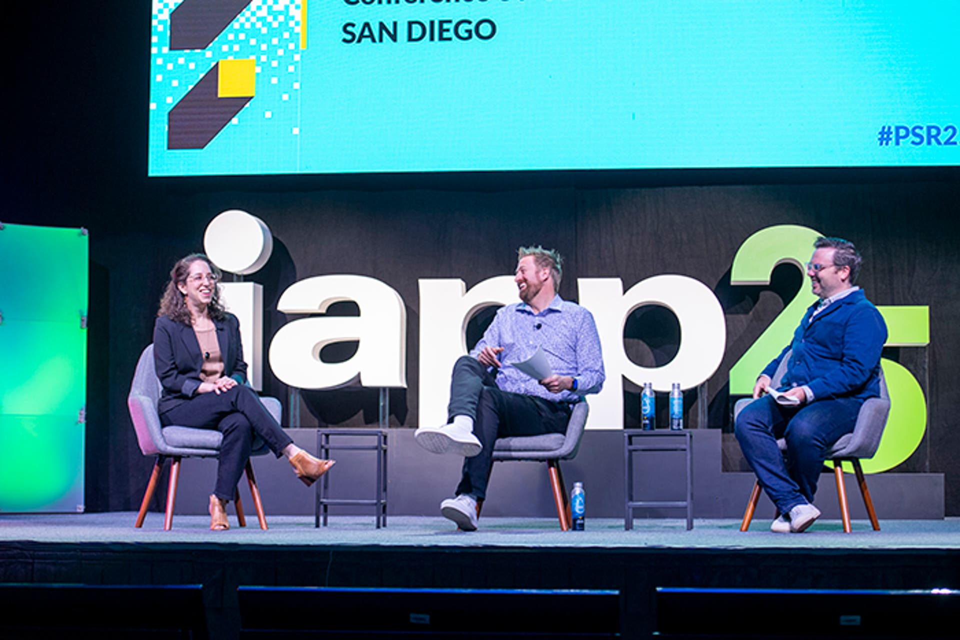 Three people seated on stage in gray chairs, participating in a panel discussion. Large “iapp25” signage and a screen displaying event details are visible behind them.