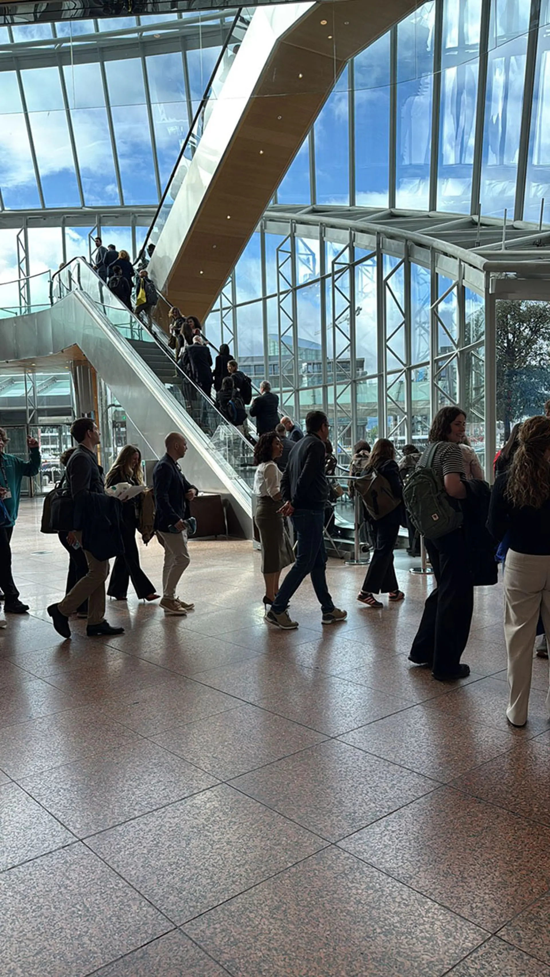 A large, modern glass atrium shows a crowd of people walking across a tiled floor while others ride an escalator that ascends diagonally through the space. The area is brightly lit by natural light coming through tall windows, and structural beams and a glass ceiling frame the open interior.