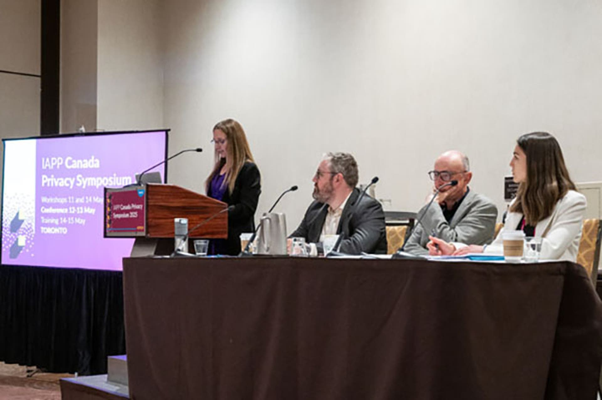 Panel discussion with four people seated at a long table covered in a black cloth. A purple screen beside the podium displays “IAPP Canada Privacy Symposium 2025.”