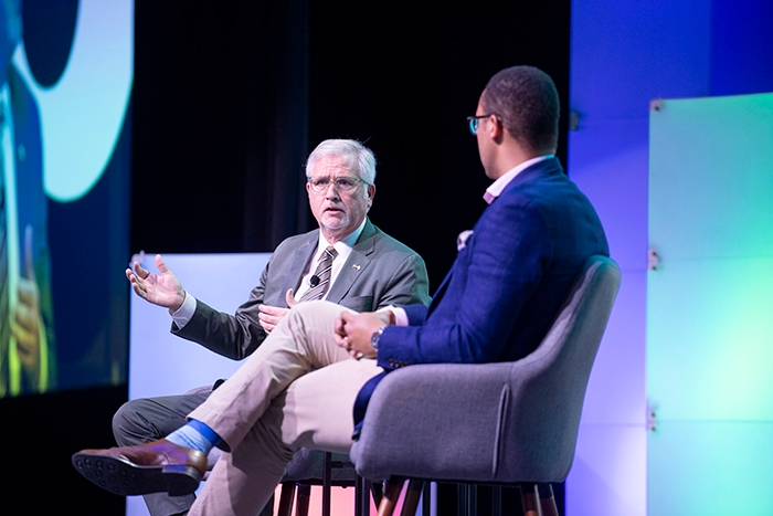 Two people seated on stage in gray chairs, conversing during a session. A large screen shows a close-up of one speaker. Colorful light panels and event branding are visible in the background.