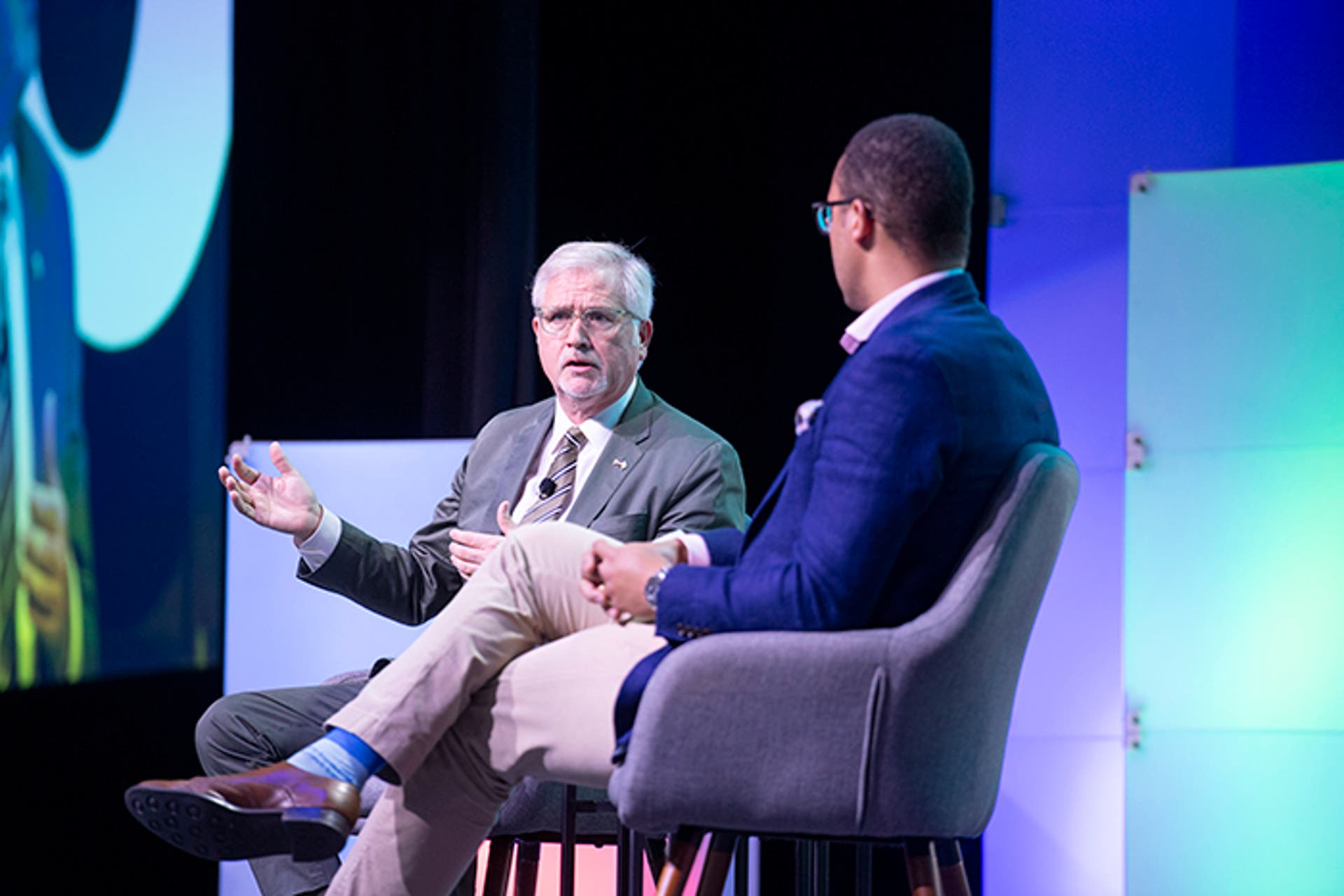 Two people seated on stage in gray chairs, conversing during a session. A large screen shows a close-up of one speaker. Colorful light panels and event branding are visible in the background.