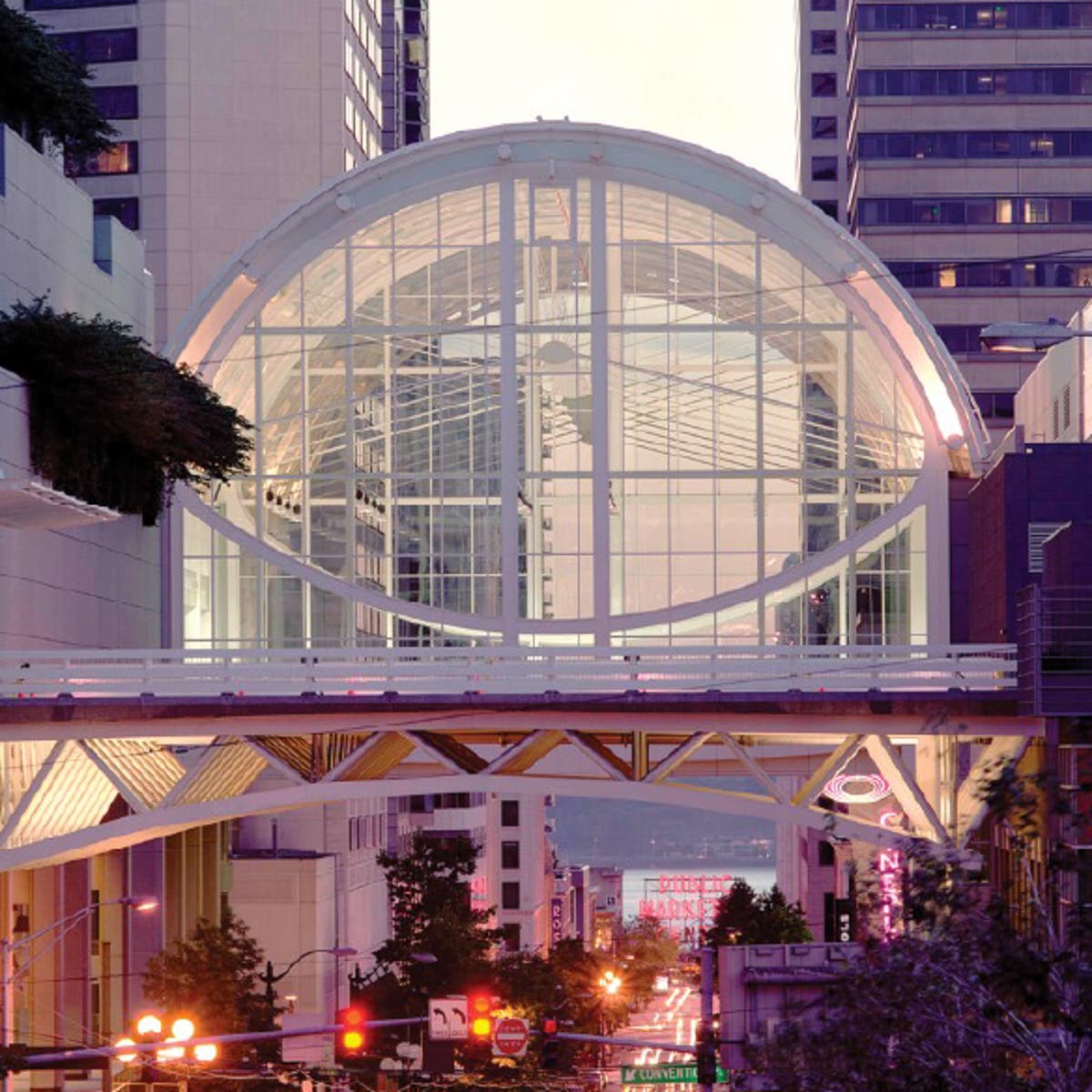 Glass-enclosed skybridge with a large circular frame and grid-like structure connecting two tall buildings in an urban setting. The bridge is illuminated with soft pink and white lighting, and city streetlights and signage are visible below.