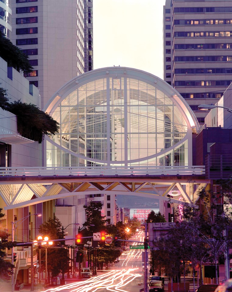 Glass-enclosed skybridge with a large circular frame and grid-like structure connecting two tall buildings in an urban setting. The bridge is illuminated with soft pink and white lighting, and city streetlights and signage are visible below.