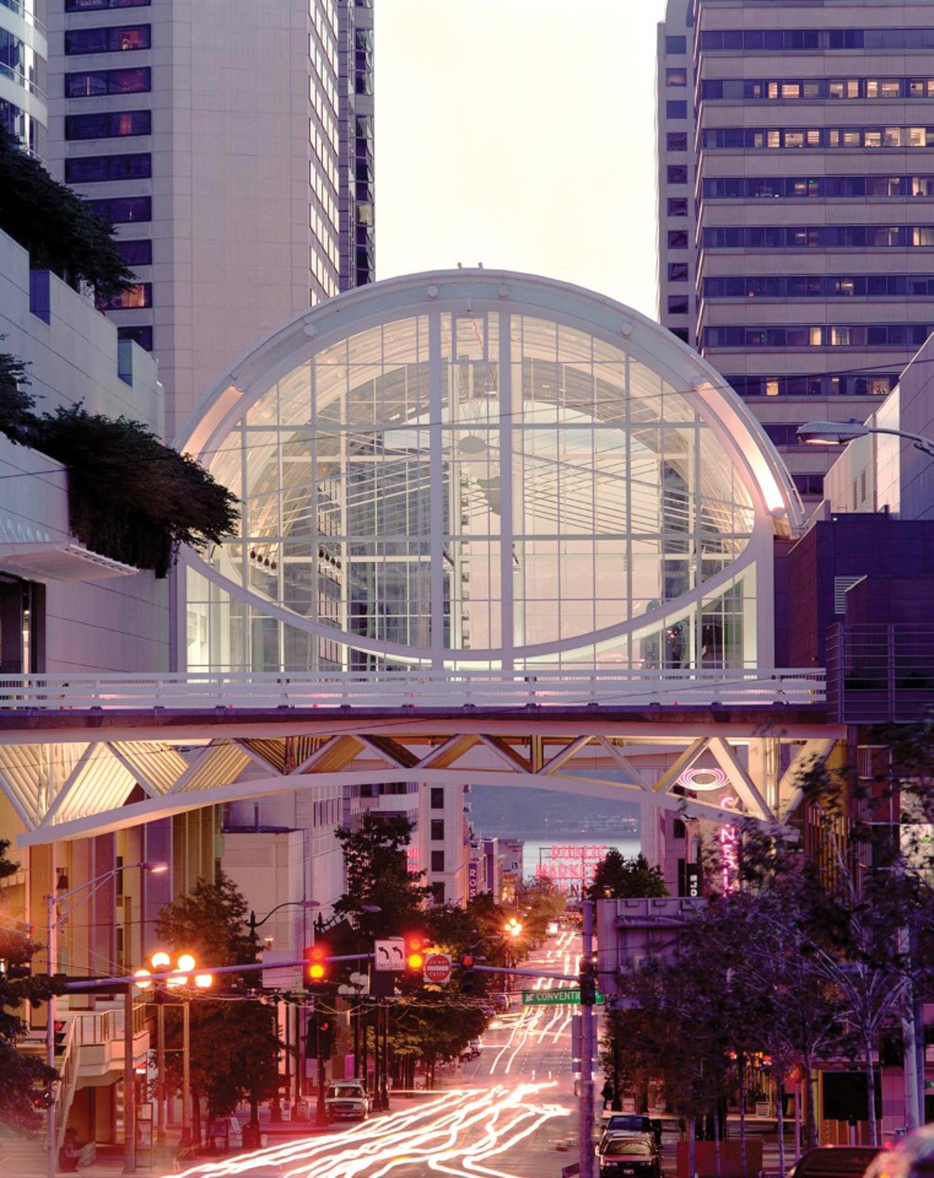 Glass-enclosed skybridge with a large circular frame and grid-like structure connecting two tall buildings in an urban setting. The bridge is illuminated with soft pink and white lighting, and city streetlights and signage are visible below.
