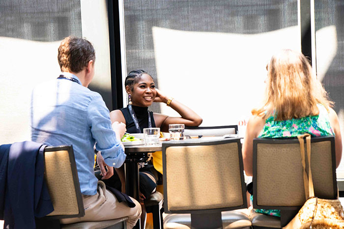 Three people seated at a small round table in a bright indoor space with large windows, engaged in conversation with drinks and notebooks on the table.