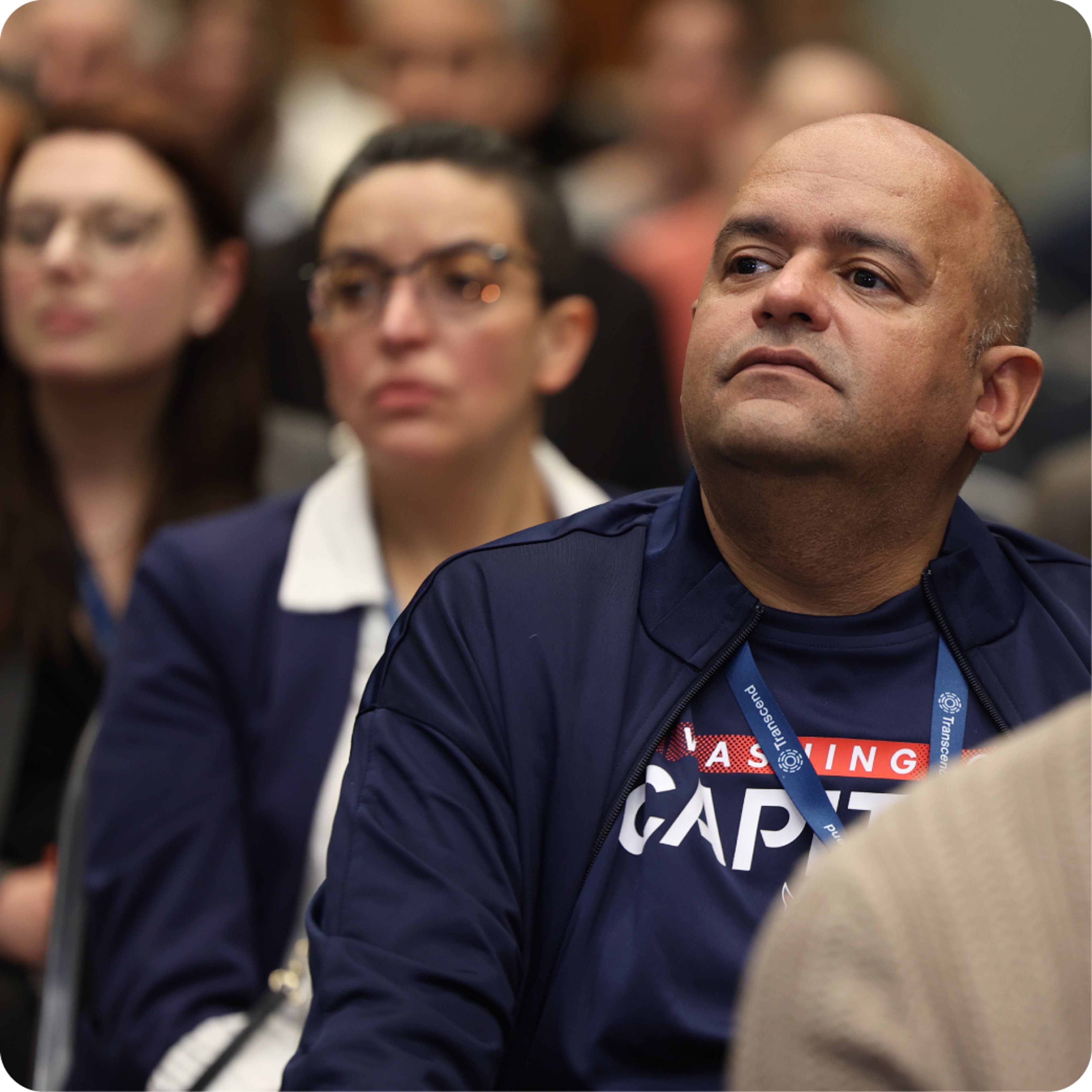 In the foreground, a man listening to a conference presentation; a woman in glasses can be seen over his shoulder.