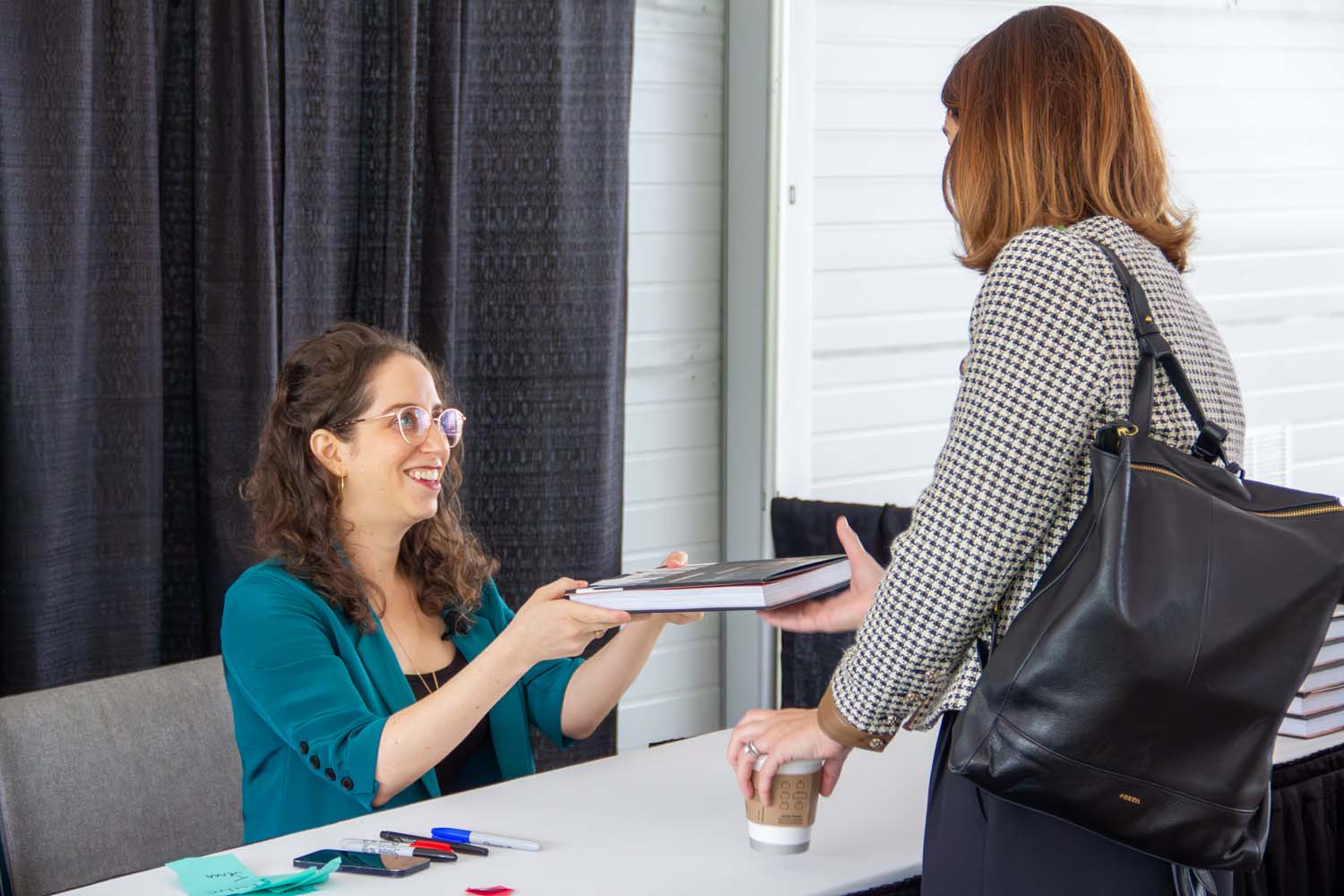 At a signing table, a speaker smiles while handing a book to an attendee, with pens, a notebook, and a coffee cup visible on the table, capturing a brief personal exchange.