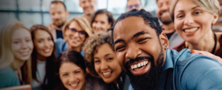 A Black man with a beard and wearing a slate blue collard shirt has an infectious smile while he hunches over slightly off center for a selfie-style photo. He's flanked by other men and women who appear slightly out-of-focus.