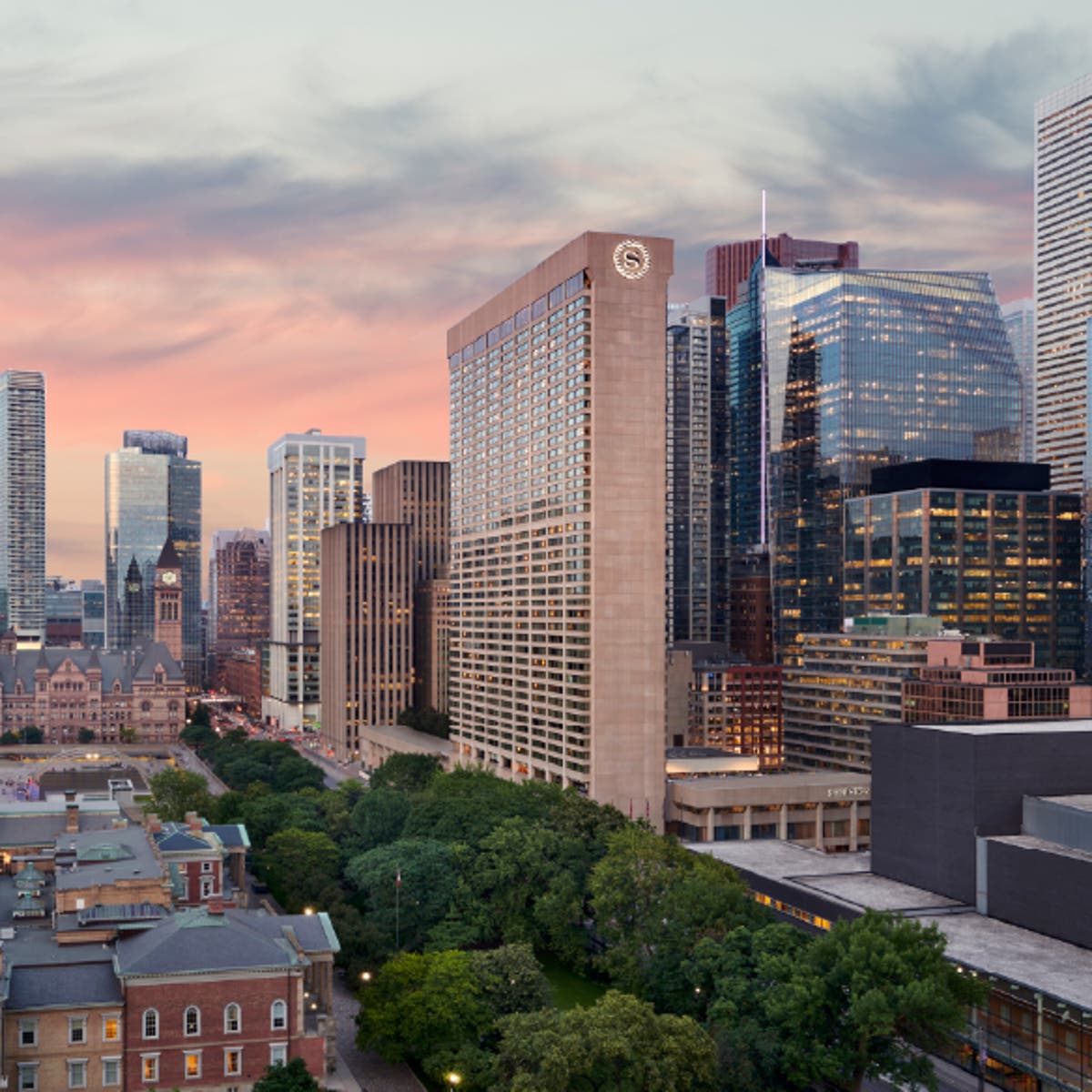 Cityscape view of downtown Toronto at sunset, featuring the Sheraton Centre hotel prominently in the foreground. Surrounding the hotel are modern glass skyscrapers and historic buildings, with green trees and a park visible below. The sky is painted with soft pink and orange hues.