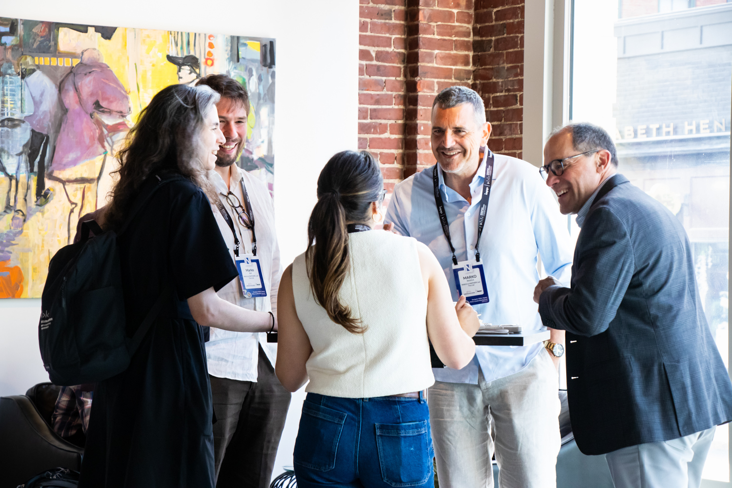 Conference attendees stand together in a sunlit networking space, conversing around a small high-top table near large windows and exposed brick walls. Event badges, artwork, and natural light contribute to the welcoming atmosphere of professional connection and conversation.