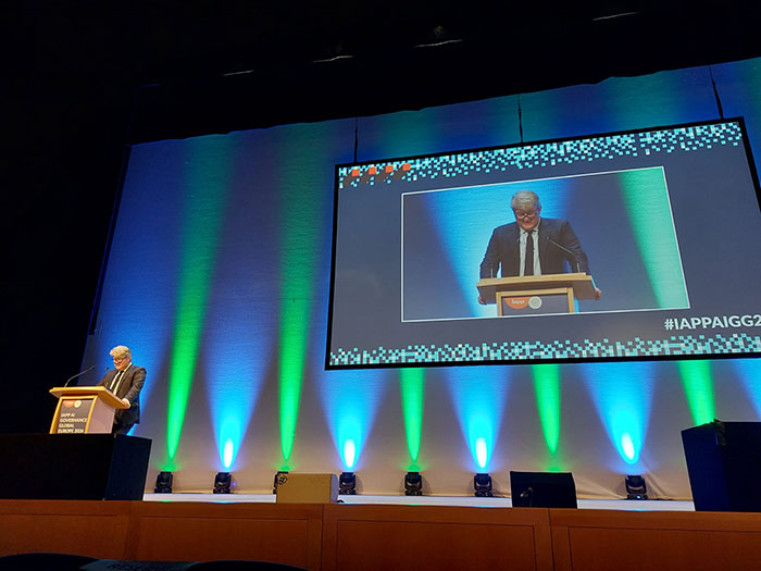 A speaker stands at a podium on a large stage while a wide screen behind them displays a live video feed of the speaker along with the event hashtag IAPPAIGG26. The stage is illuminated with blue and green vertical lighting, and the setting resembles a large conference auditorium.