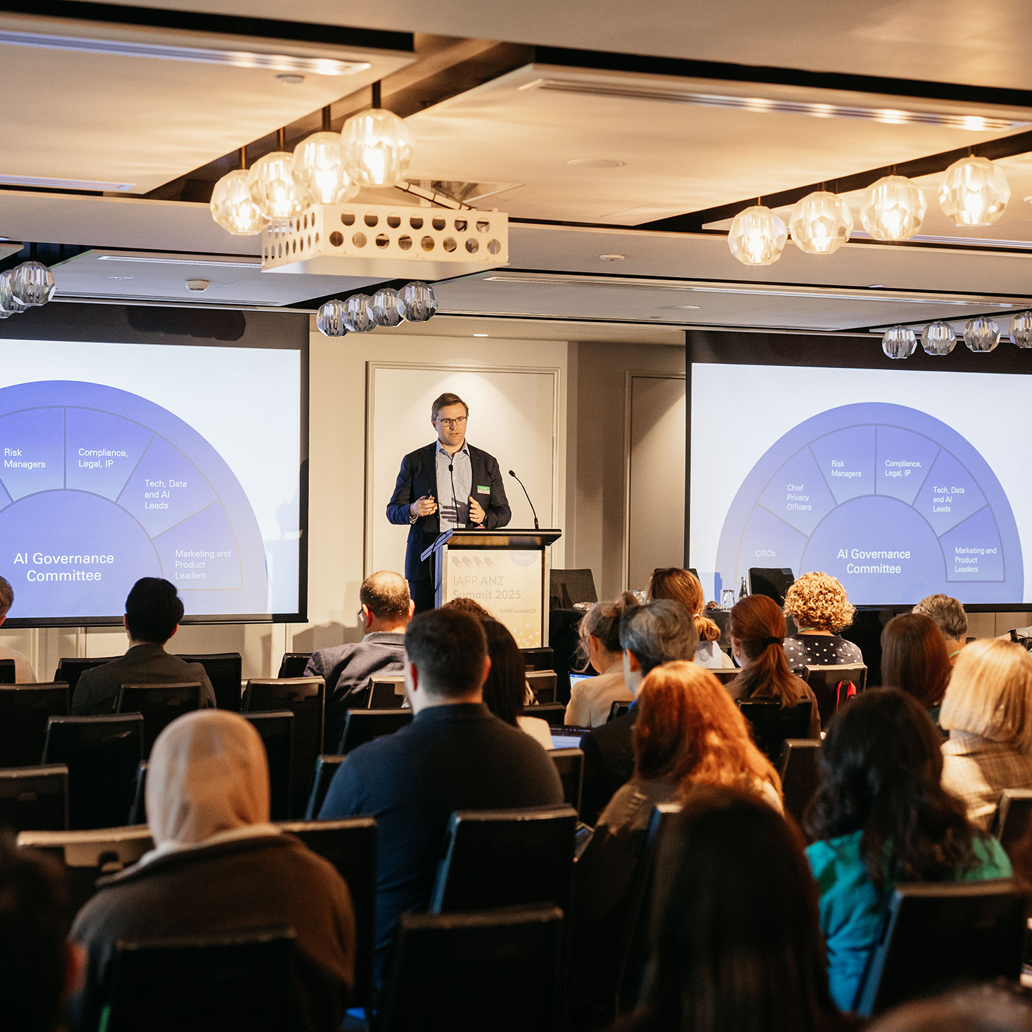 Presenter at a podium in a conference room with two screens showing a circular AI governance framework diagram.