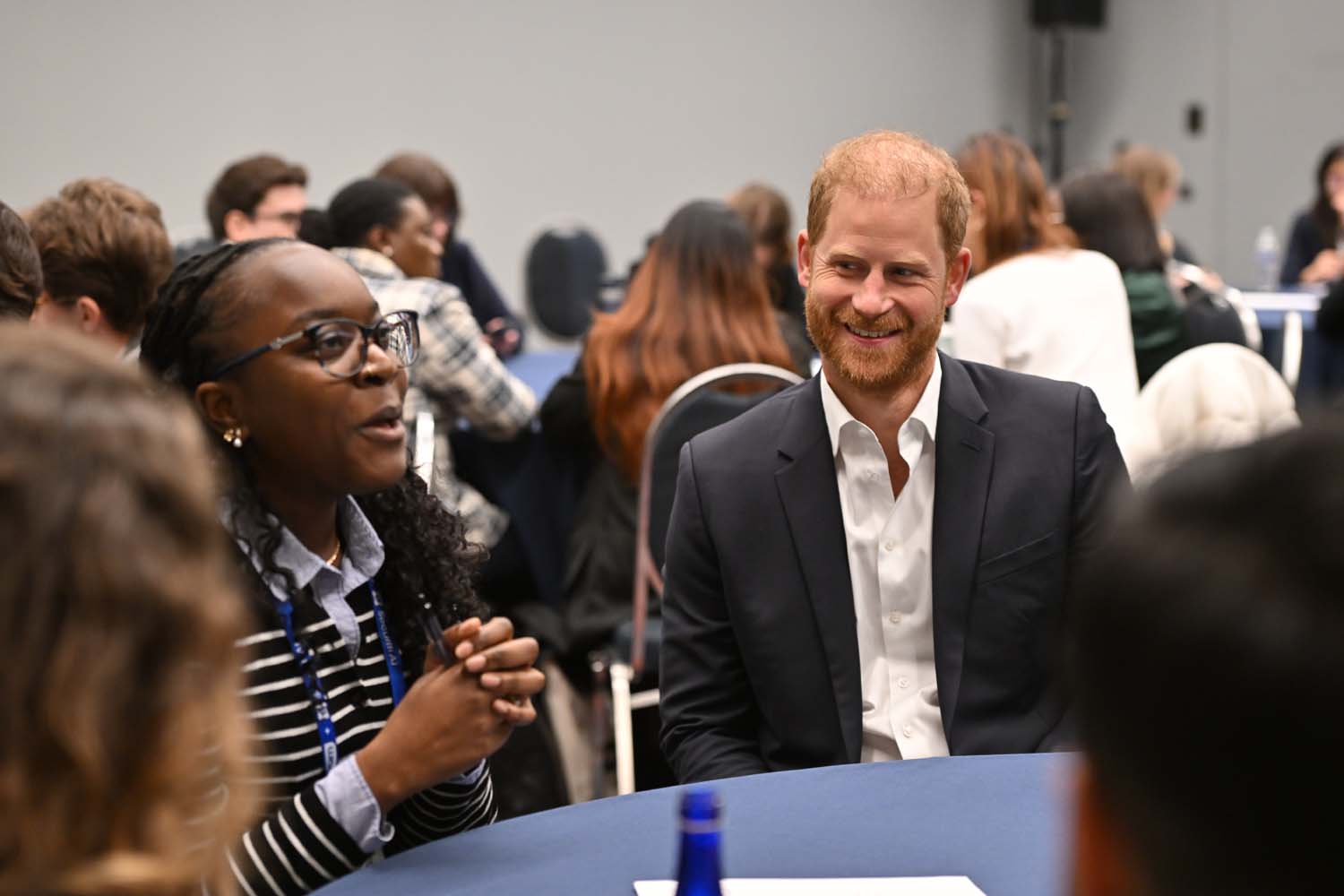 Prince Harry and several conference participants sit around a round table in a meeting room, engaged in discussion during a networking or breakout session while other tables fill the background.